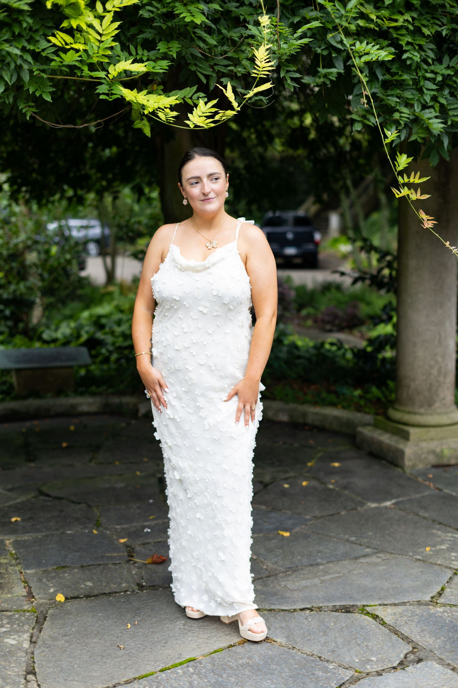 Woman in white floral textured gown poses in lush garden setting on stone patio.