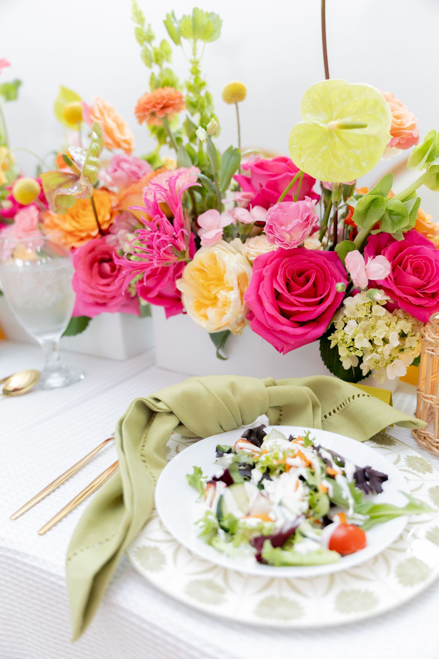 Elegant table setting with fresh salad, green napkin, gold cutlery, and vibrant pink, orange floral centerpiece.