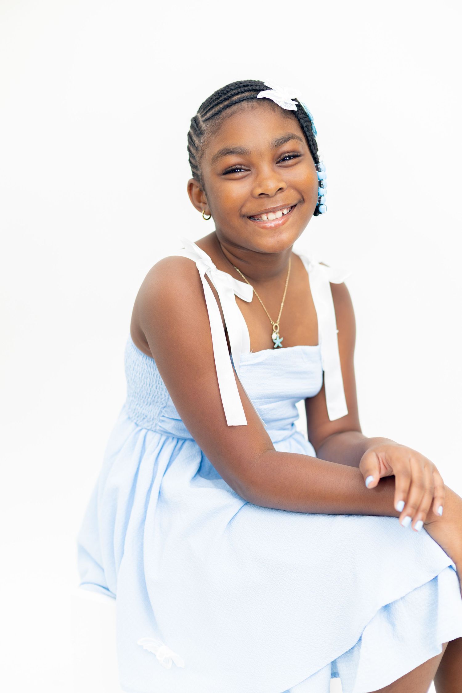 Young Black girl in light blue dress with white bow straps, braided hair, smiling confidently against white background.