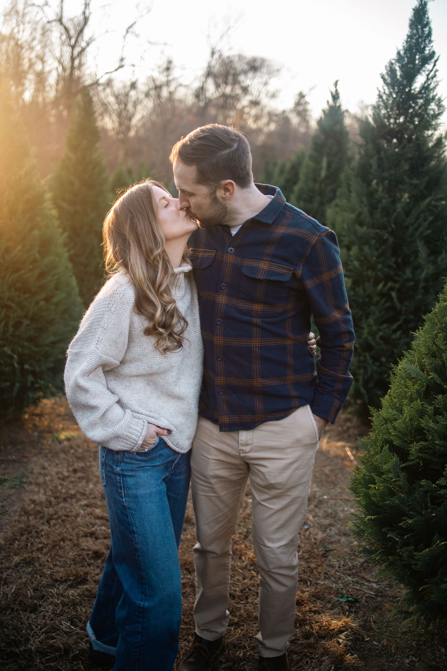 Romantic couple sharing intimate moments among evergreen trees at sunset in a Christmas tree farm.