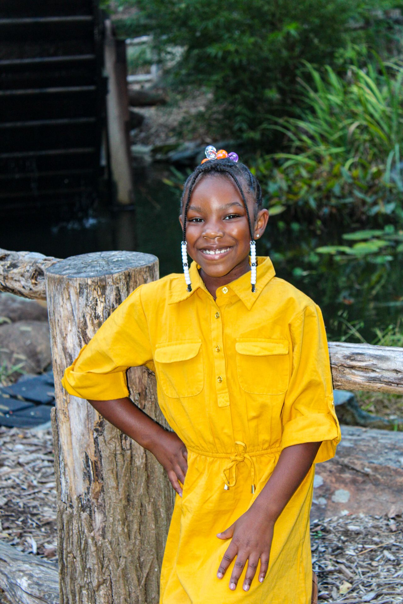 Child posing in a bright yellow dress next to tree stumps in an outdoor setting.