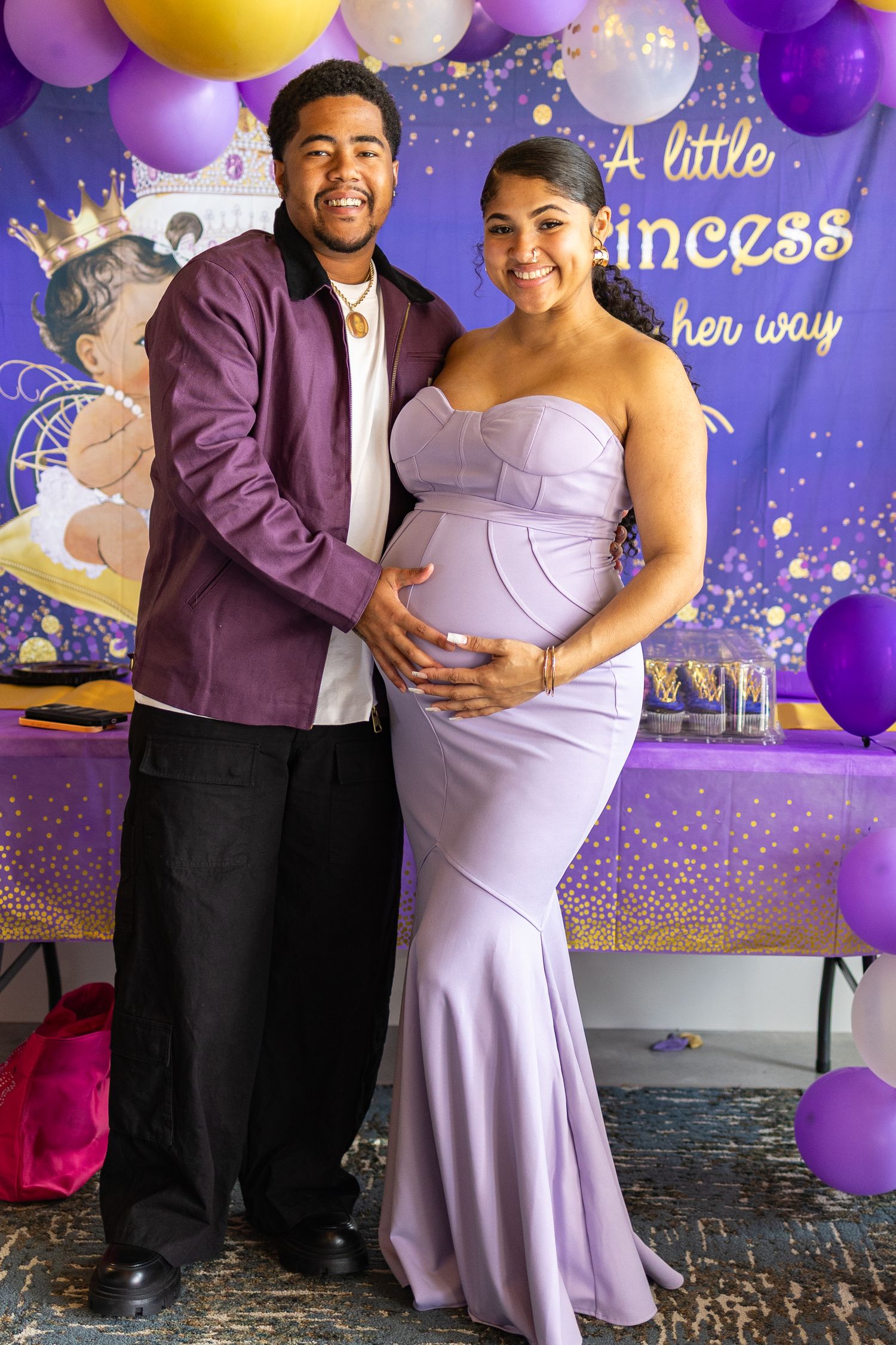 Two people stand together at a celebration with purple and gold balloons and a decorated backdrop.