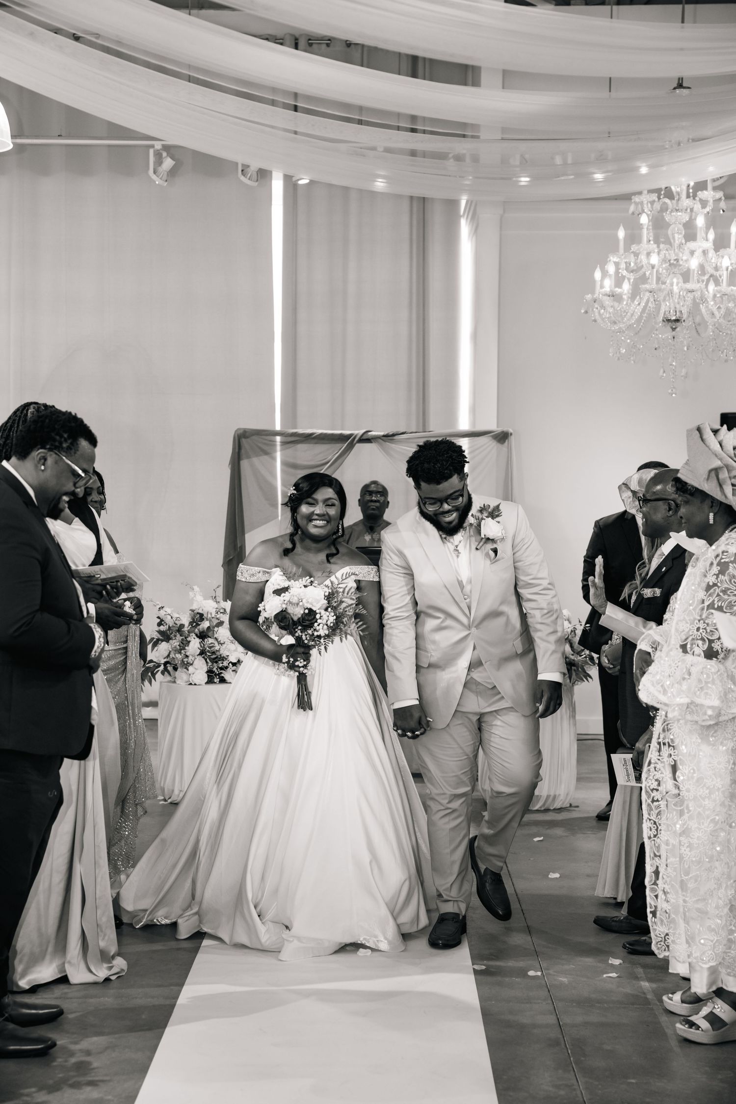 Wedding celebration with guests dancing and mingling in an elegant white reception hall with chandeliers.