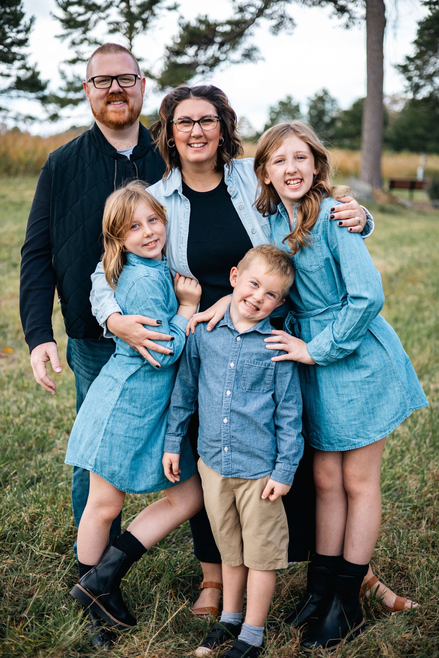 A family wearing coordinating denim and blue outfits poses playfully together in a grassy outdoor setting.