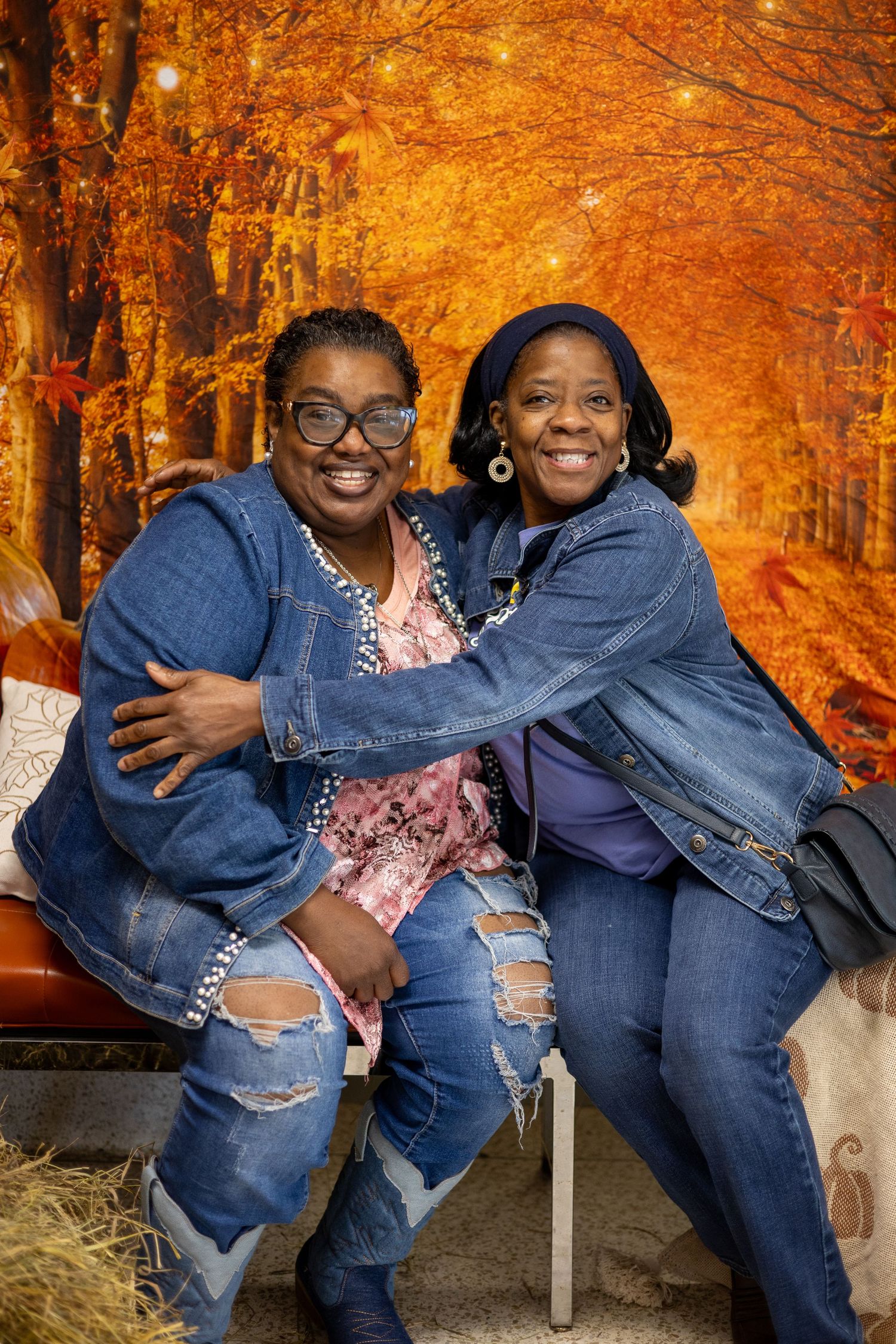 Two people in denim outfits share joyful laughter while sitting on a bench surrounded by vibrant autumn foliage.