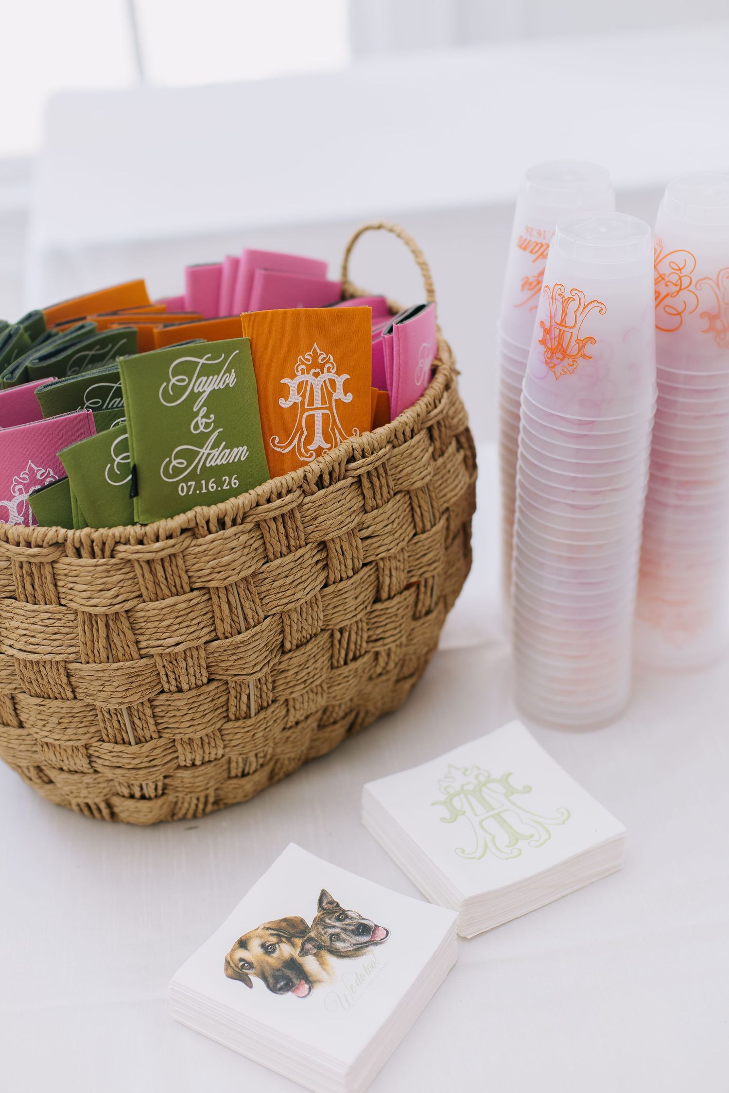Woven basket with colorful wedding favor packets for Taylor & Adam, stacked frosted cups, and monogrammed napkins on white table.