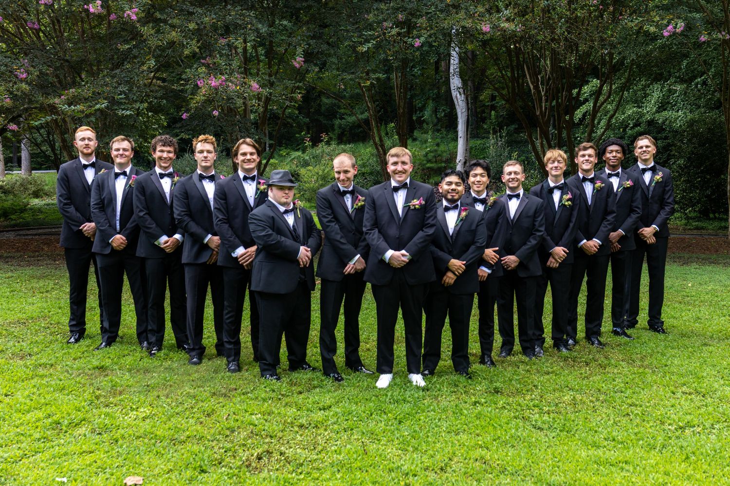 Large groomsmen group in black tuxedos and bow ties posing outdoors on green lawn with lush trees behind.