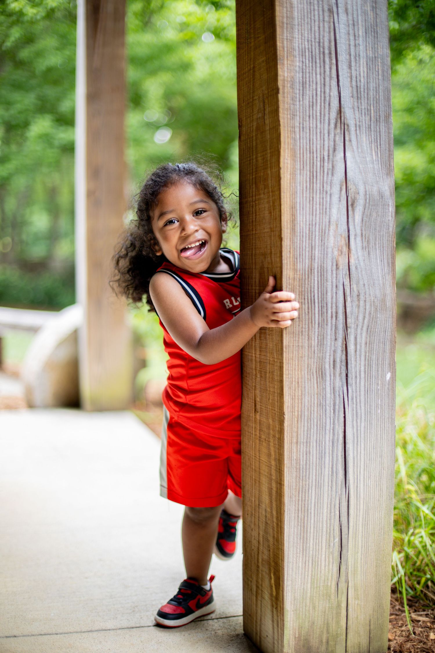 Happy toddler girl in red outfit hugging a wooden post outdoors, smiling joyfully in a park setting.
