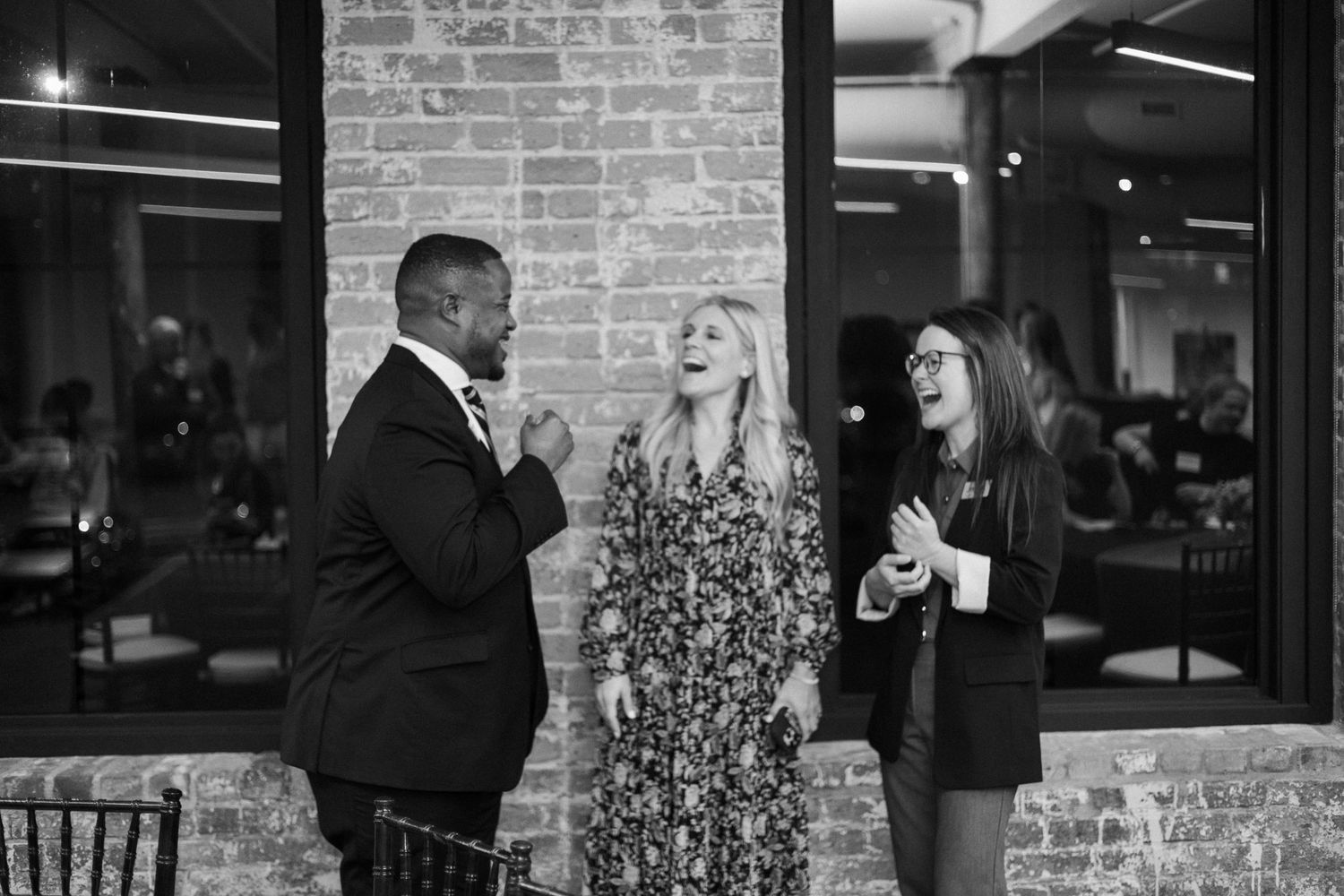 Three people laugh and converse outdoors near a brick wall at a social gathering.