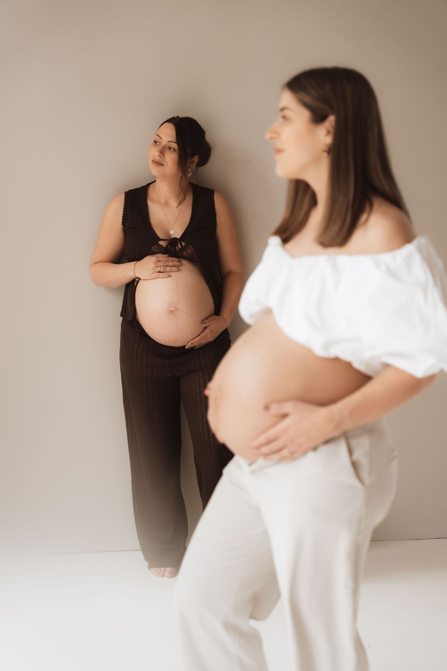 Two pregnant women cradle their baby bumps in a studio, one in black outfit and one in white off-shoulder top.