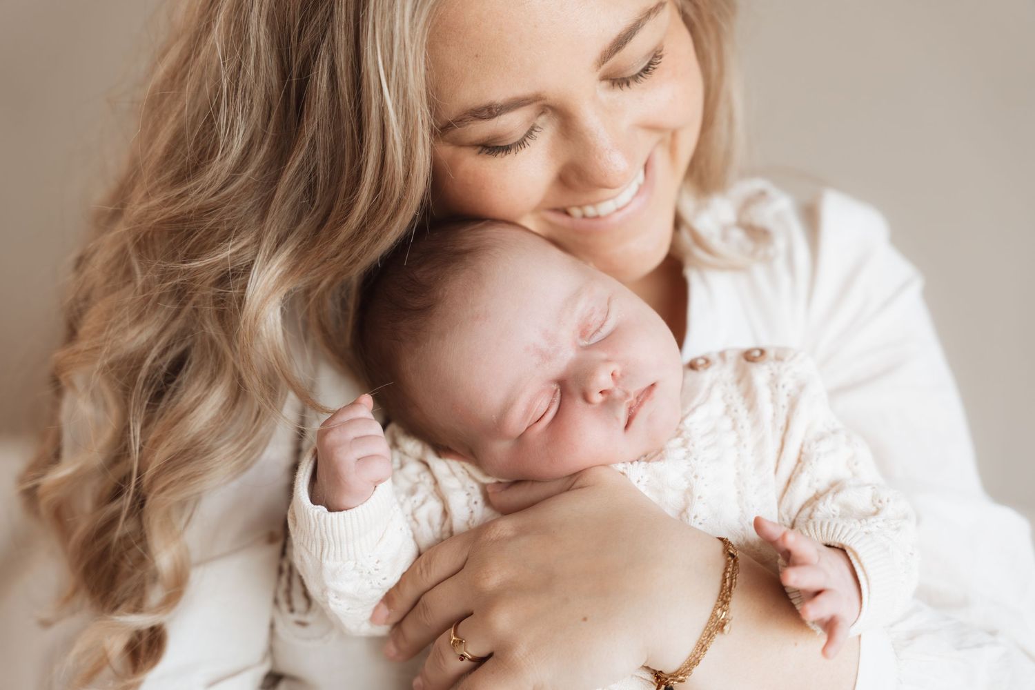 Smiling mother with long blonde hair gently holds sleeping newborn baby dressed in white knit outfit.