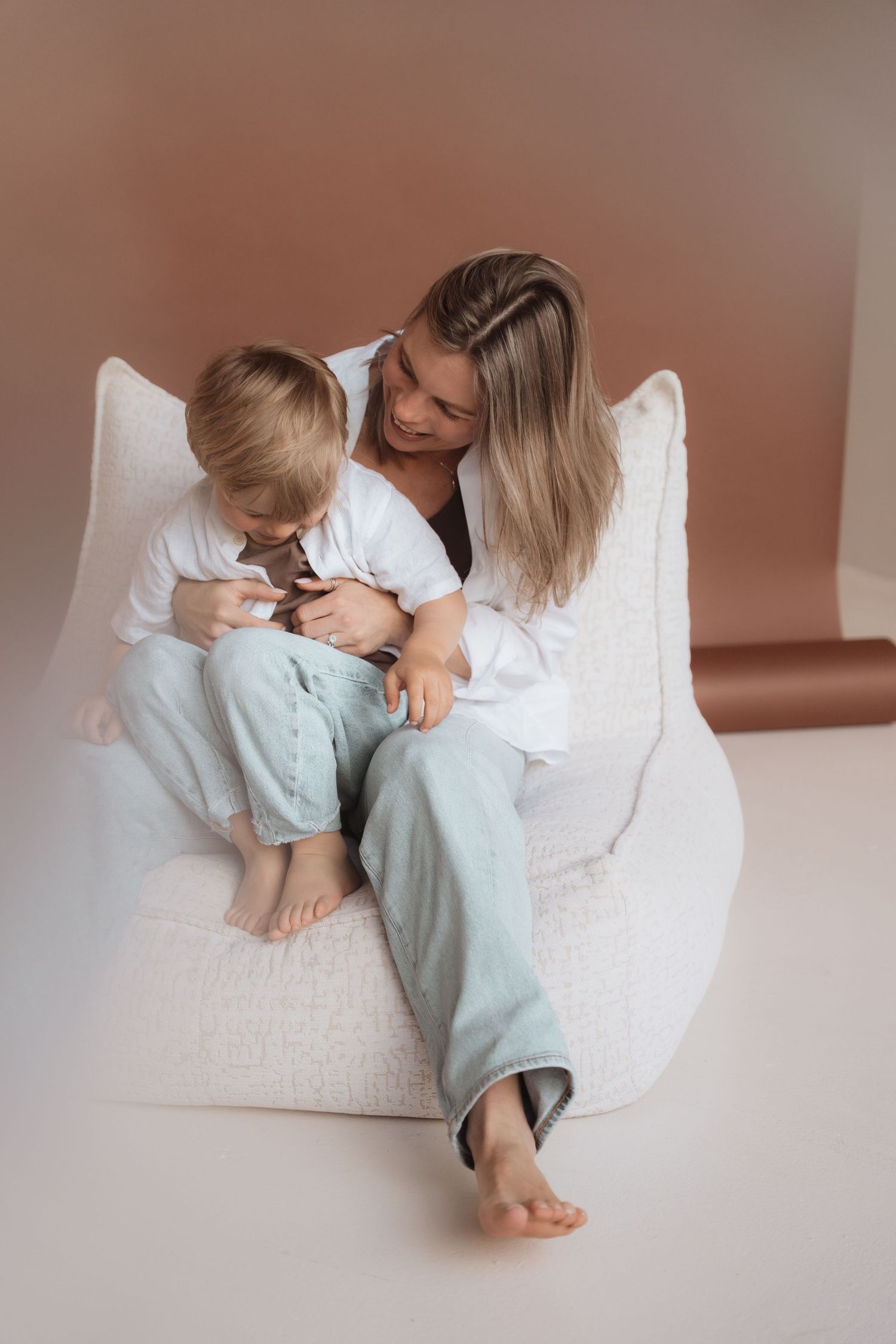 Mother and young son sitting together on white bean bag chair, both wearing casual white shirts and jeans.