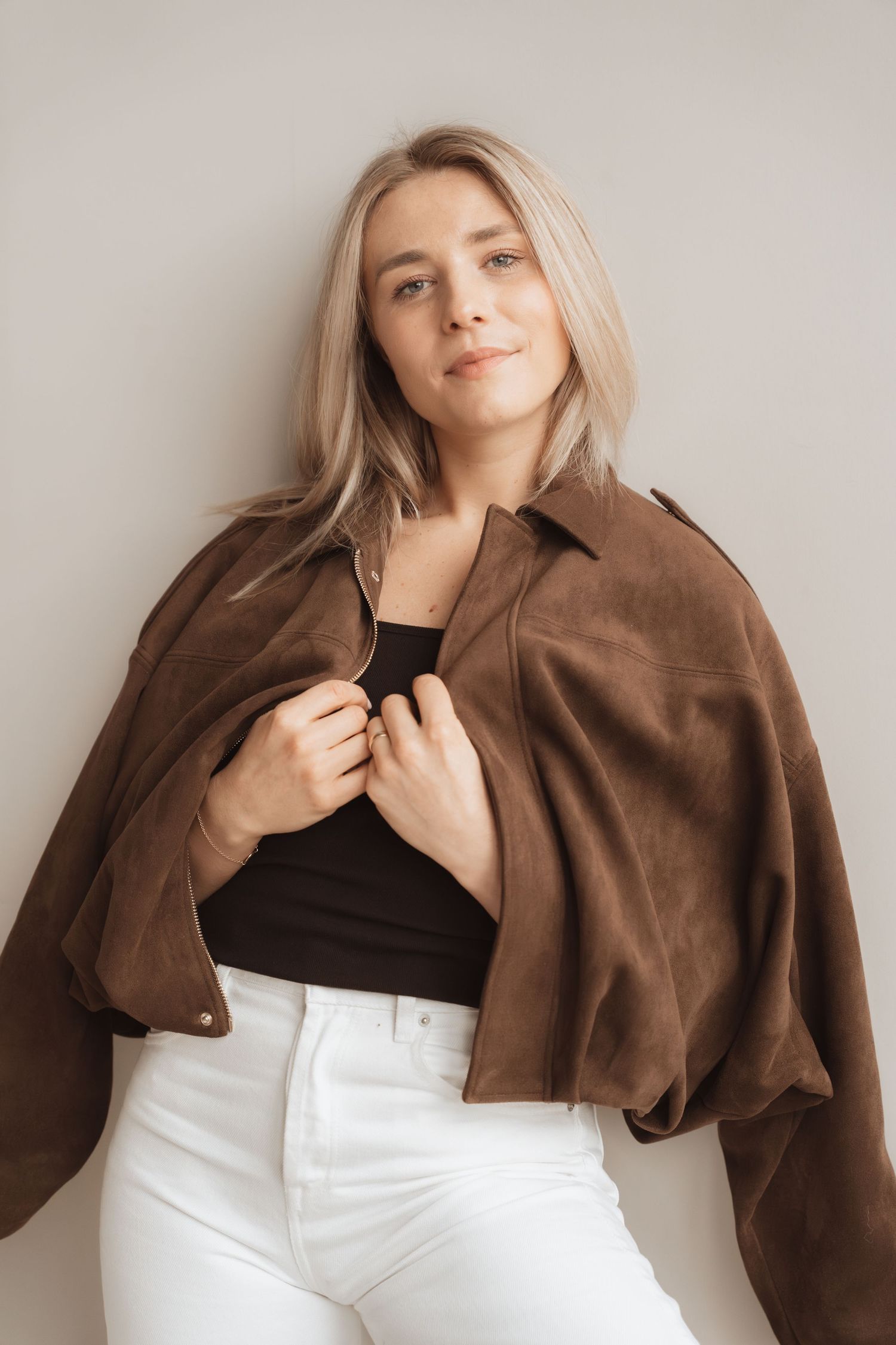 Blonde woman posing in brown suede jacket over black crop top with white jeans against neutral background.