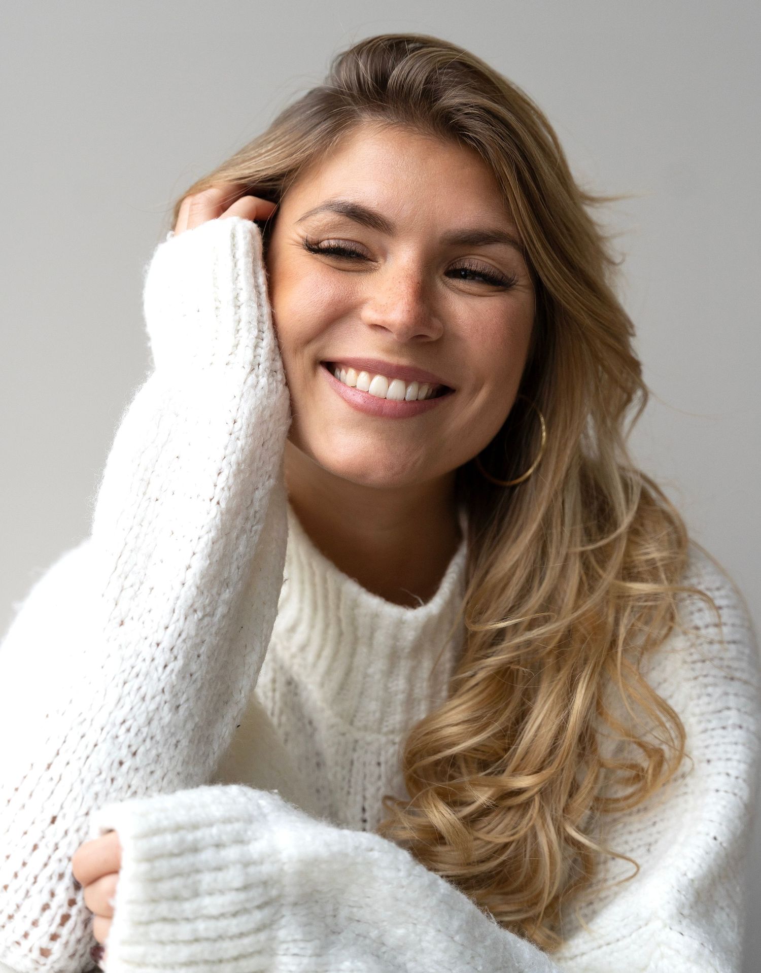 Woman in cozy white knit sweater smiles warmly while posing against light background.