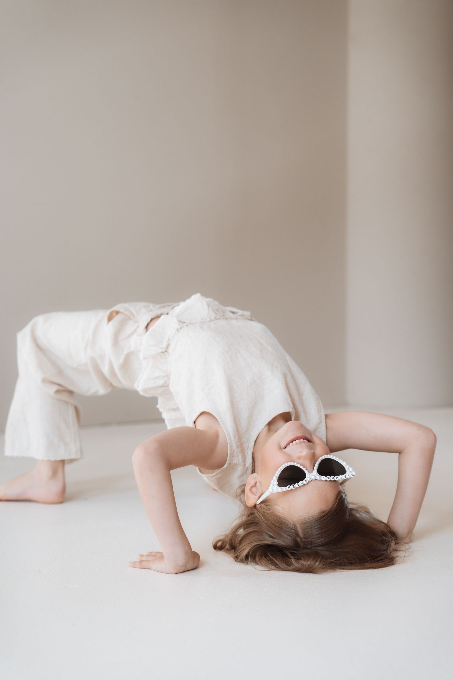 Young girl in white outfit and cat-eye sunglasses doing a backbend on a light studio floor.