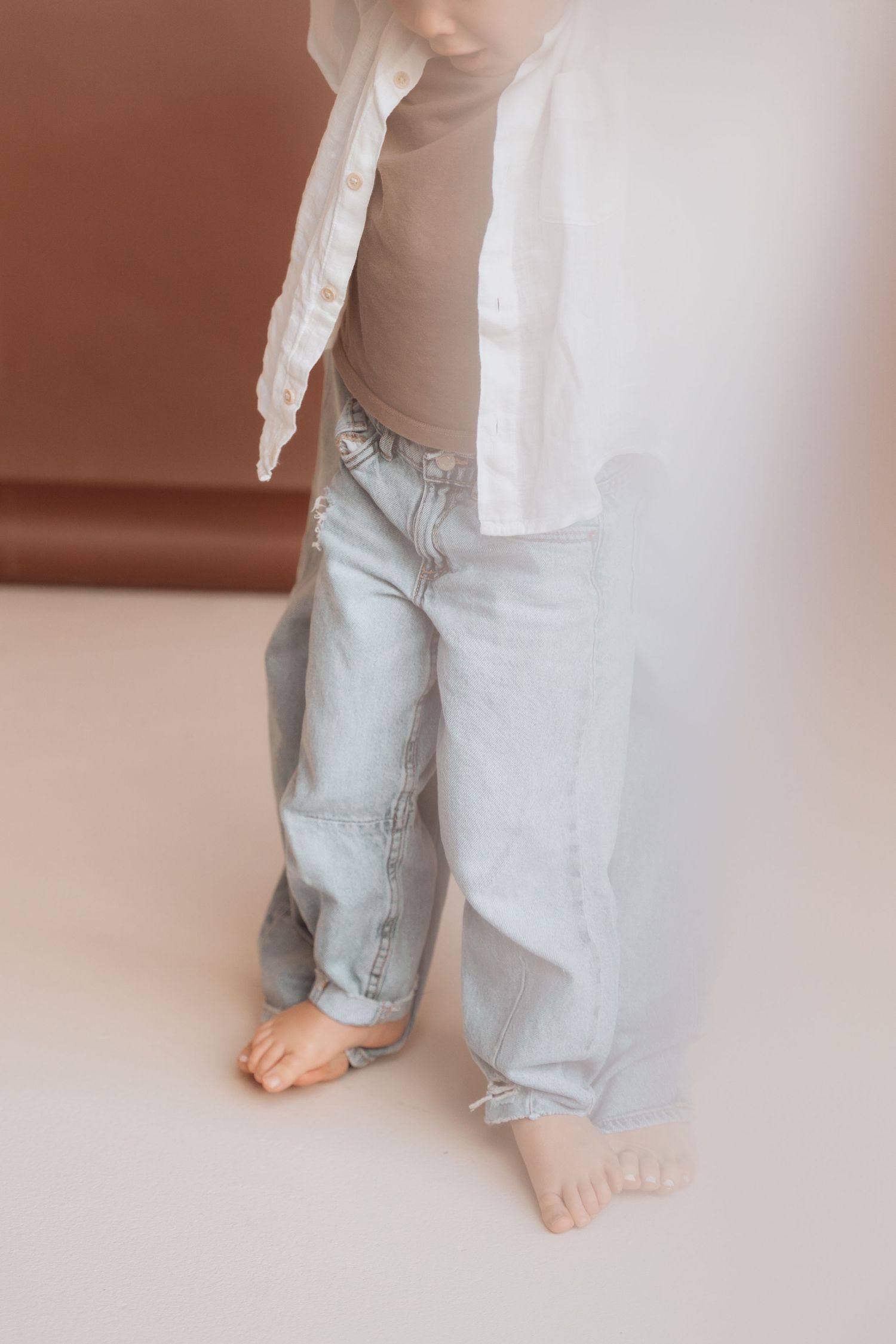 Toddler standing barefoot in light blue jeans and white linen shirt over beige top on white floor.