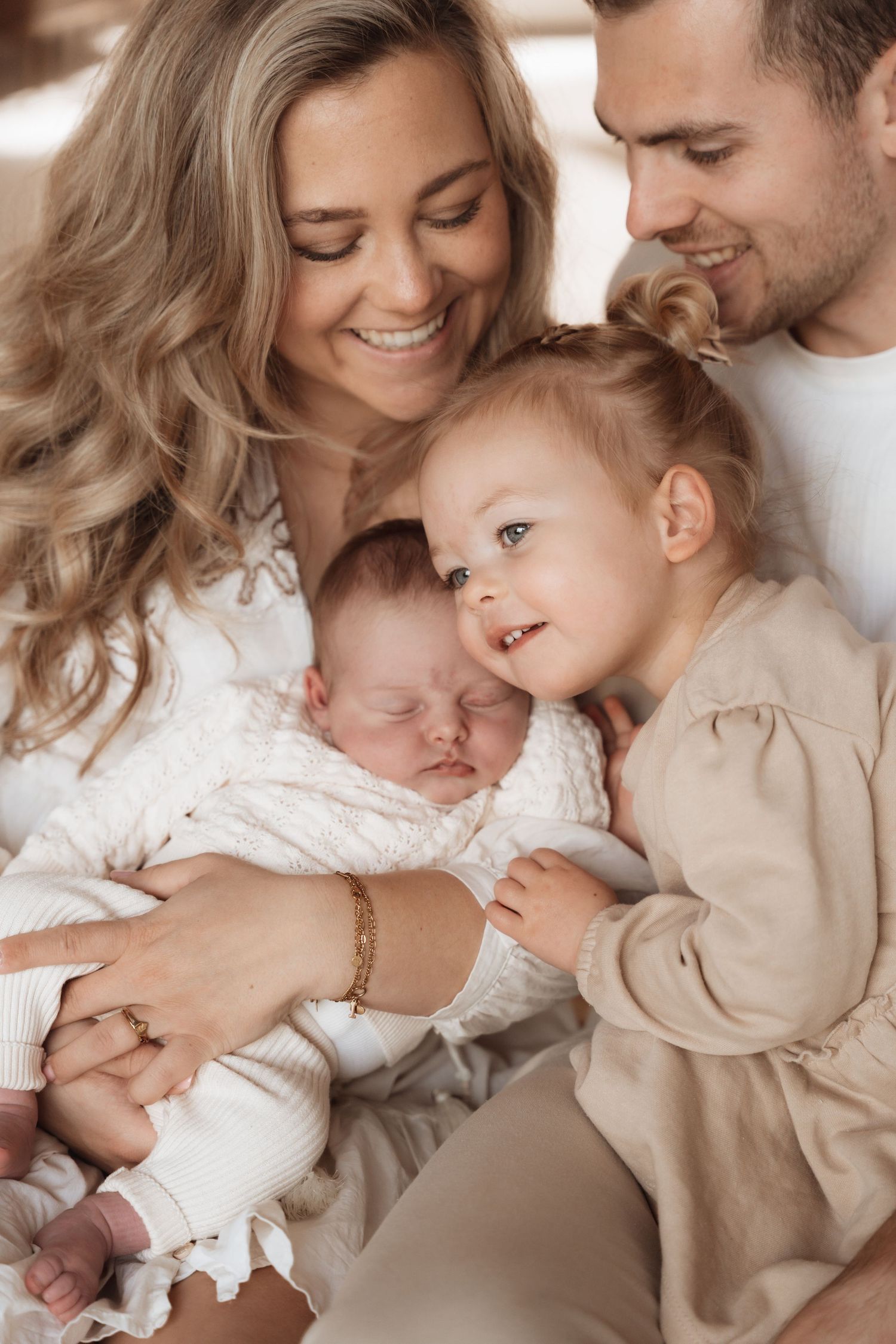 Happy family with parents smiling at newborn baby held by mother, with toddler girl leaning in affectionately.