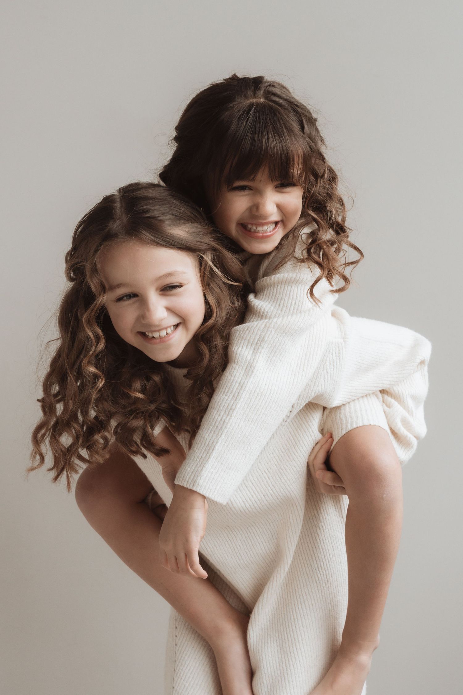 Two smiling girls with curly brown hair wearing white knit outfits, one giving the other a piggyback ride.