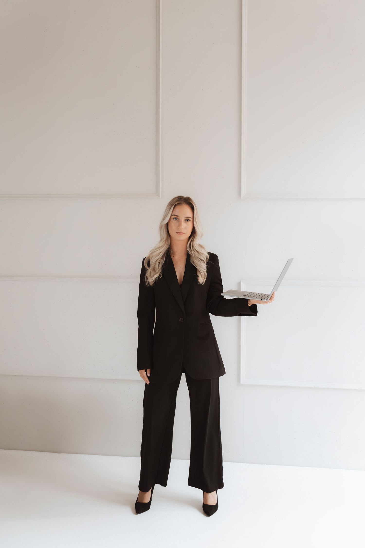 Professional woman in black suit holding laptop, standing confidently against a white wall in a modern office setting.