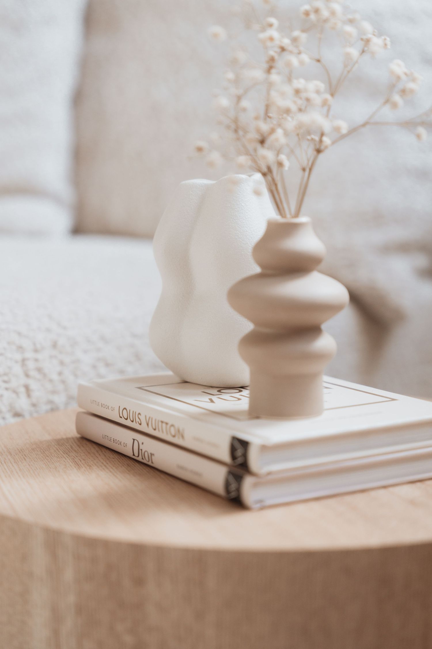A white ceramic vase with dried baby's breath flowers sits atop a coffee table book in a minimalist neutral setting.