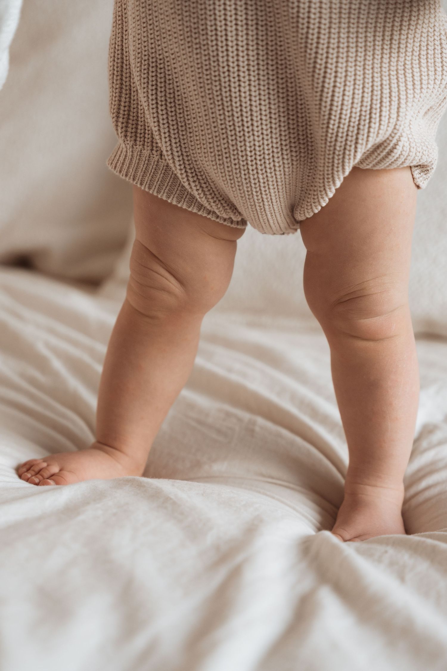 Close-up of chubby baby legs standing on white bedding, wearing a beige knit romper.