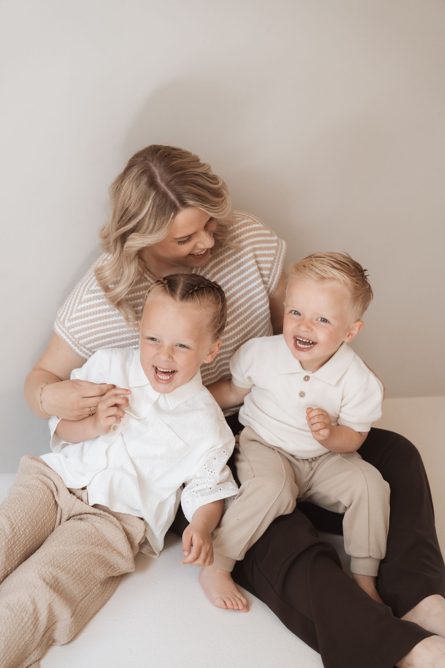 A joyful mother with blonde hair laughs with her two young sons in matching neutral outfits during a family photo session.
