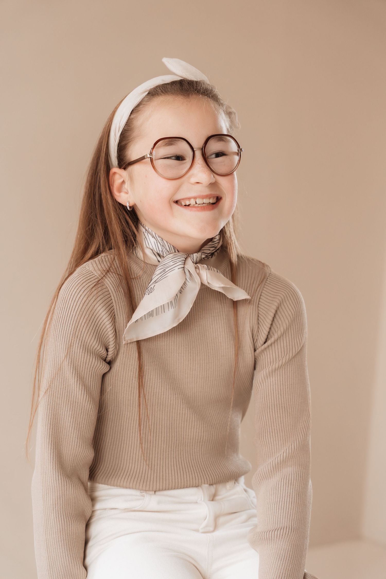 Smiling girl wearing round glasses, beige sweater, white scarf, and hair bow poses against a neutral background.