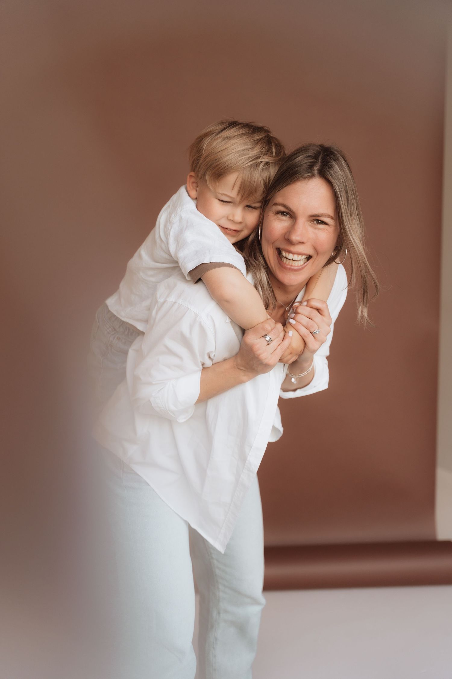 A smiling woman in white gives a young boy a piggyback ride against a warm brown background.