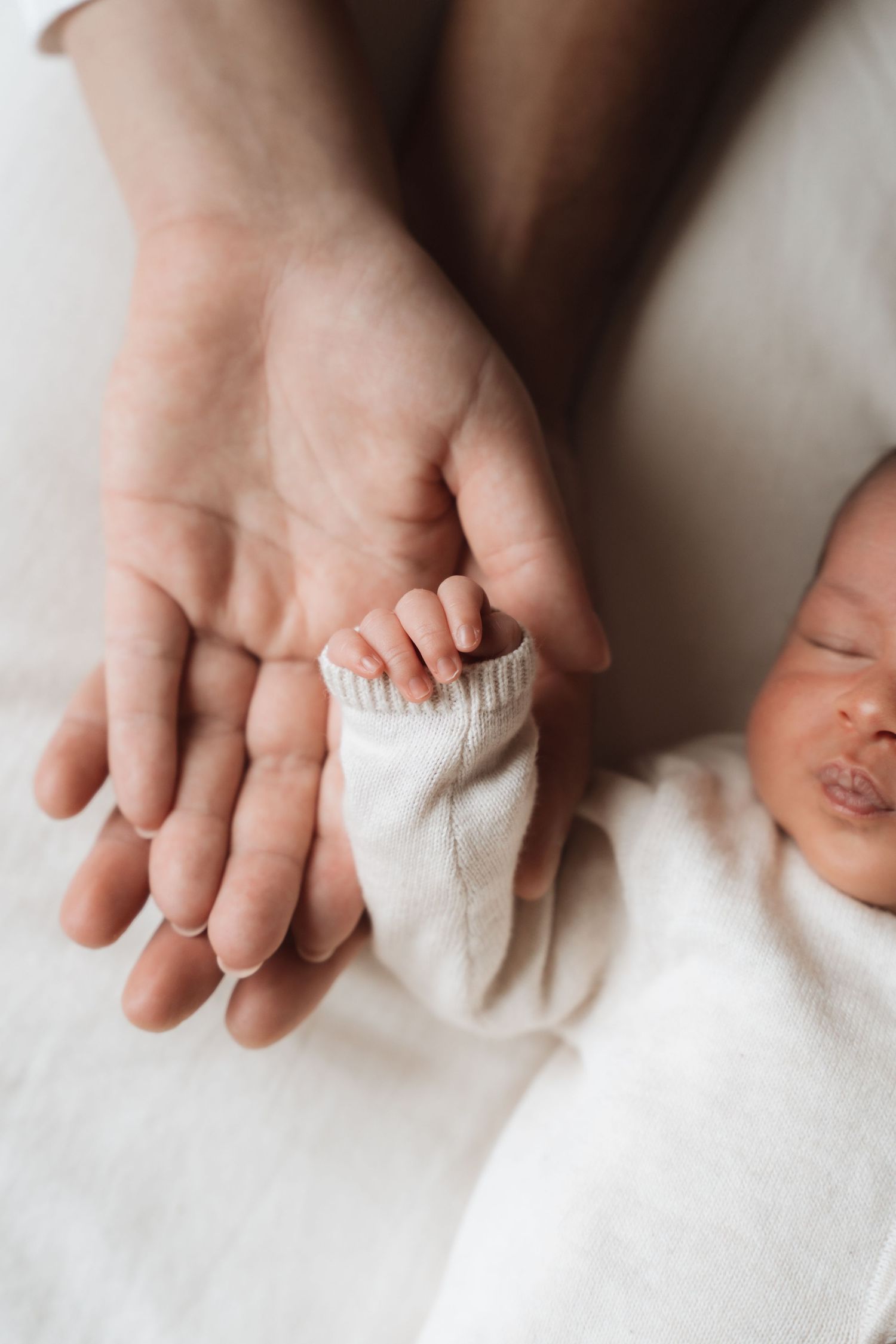 A newborn baby grasping an adult's finger with tiny hands, wrapped in a white blanket, in a tender bonding moment.