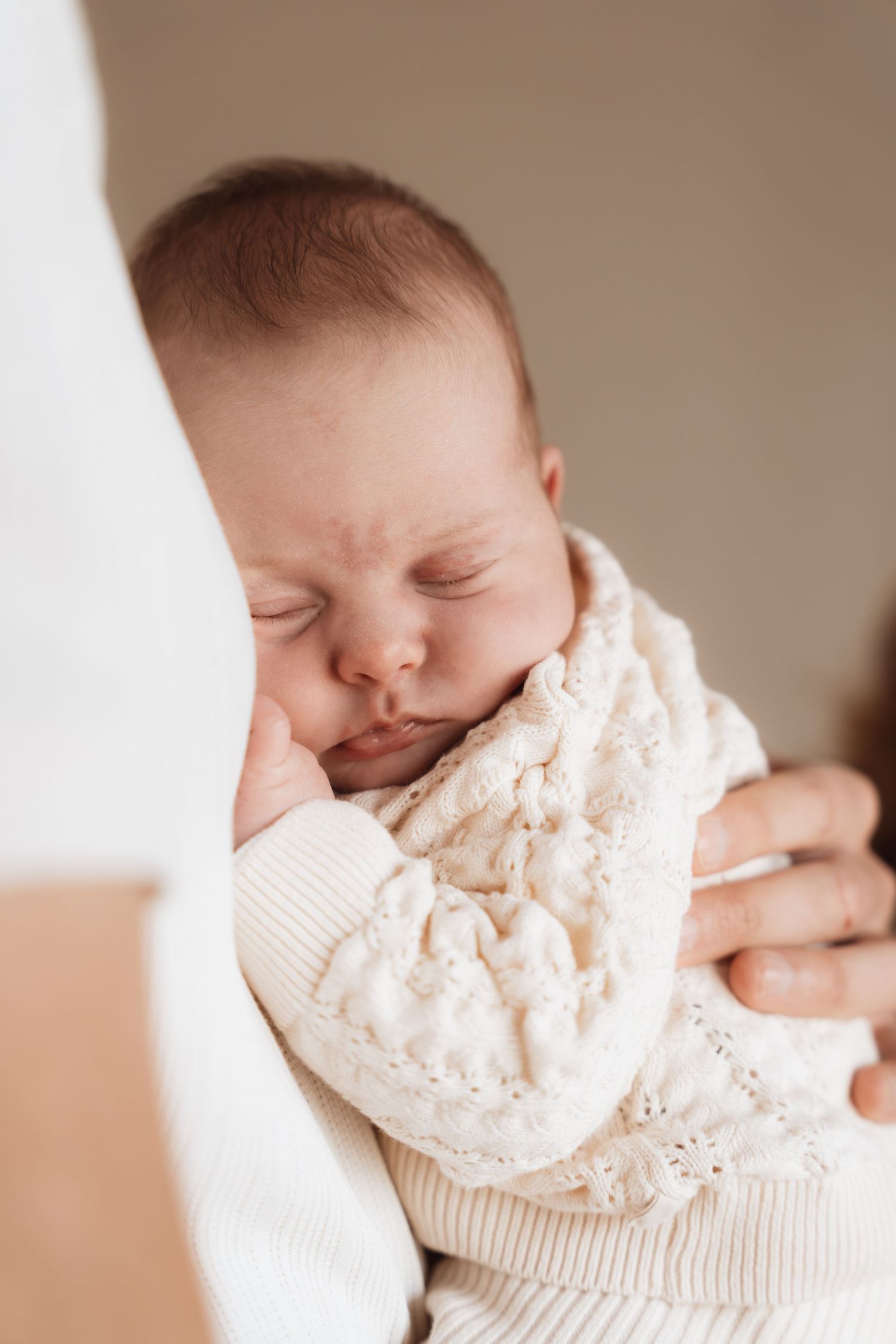 Newborn baby sleeping peacefully in a white knit outfit, cradled in a parent's arms.
