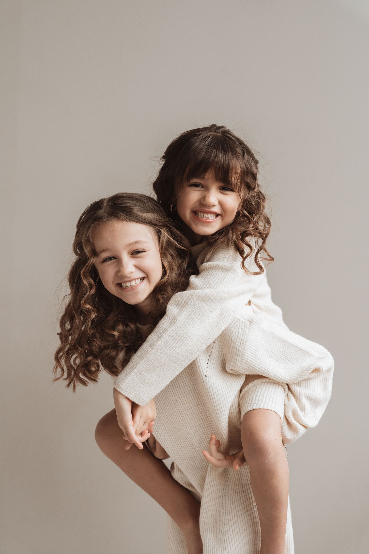 Two young girls in white knit dresses laugh joyfully as one gives the other a piggyback ride against a neutral background.