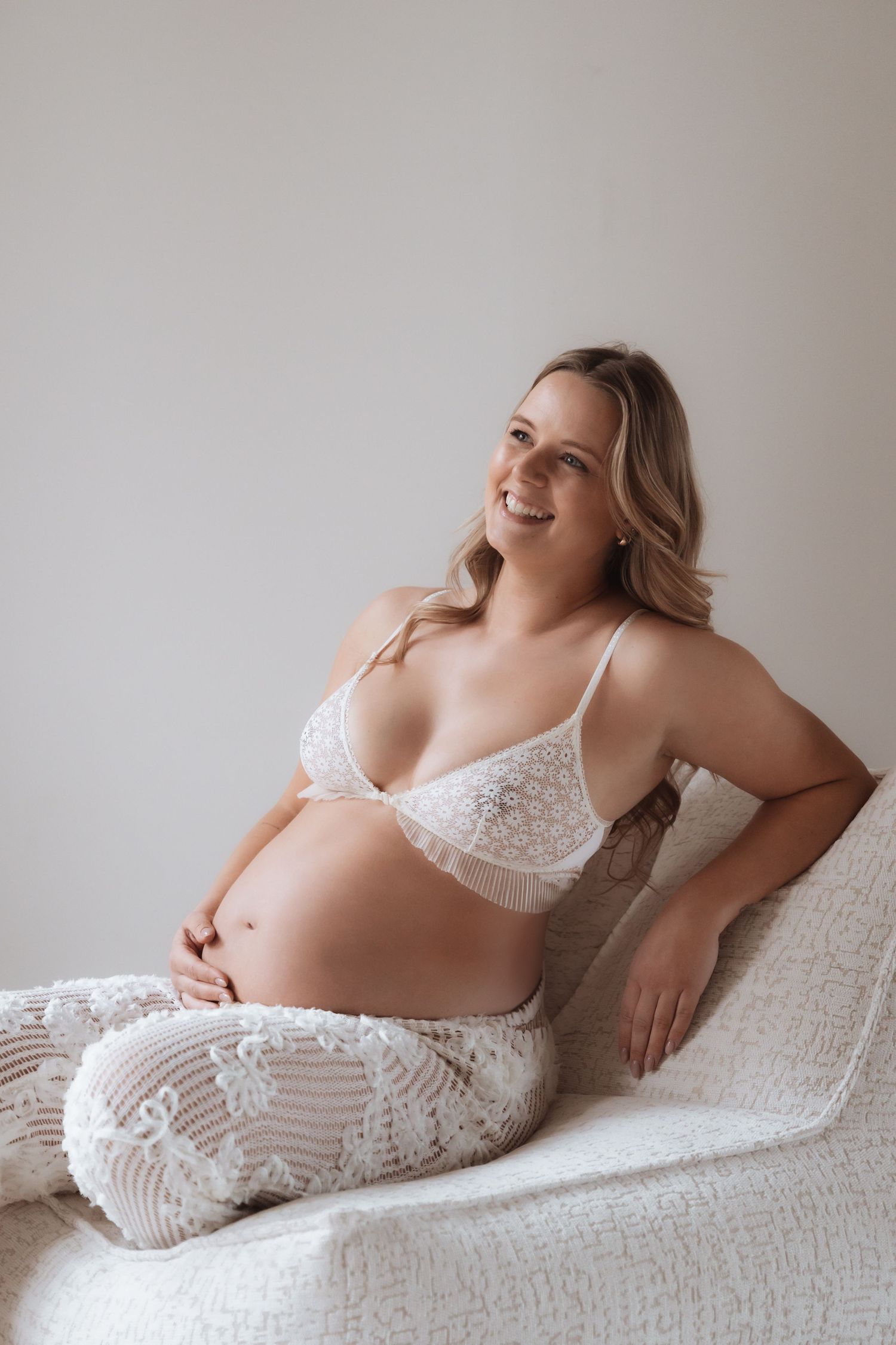 A maternity photo series showing an expectant mother in white lace lingerie reclining on light bedding in soft natural light.