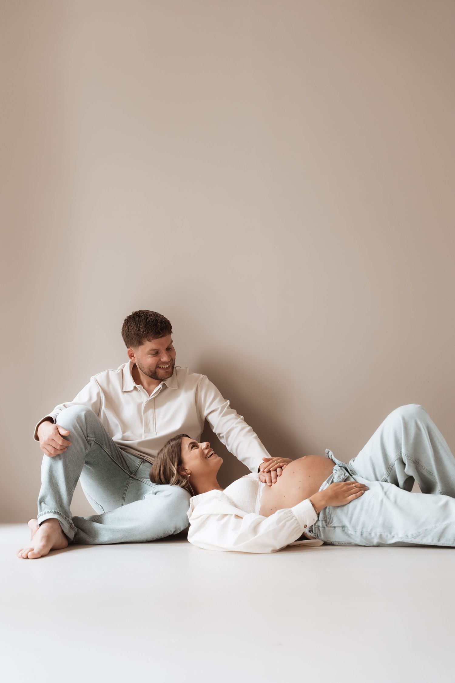 Smiling couple sitting on floor, pregnant woman lying down with exposed baby bump, wearing white shirts and light blue jeans.