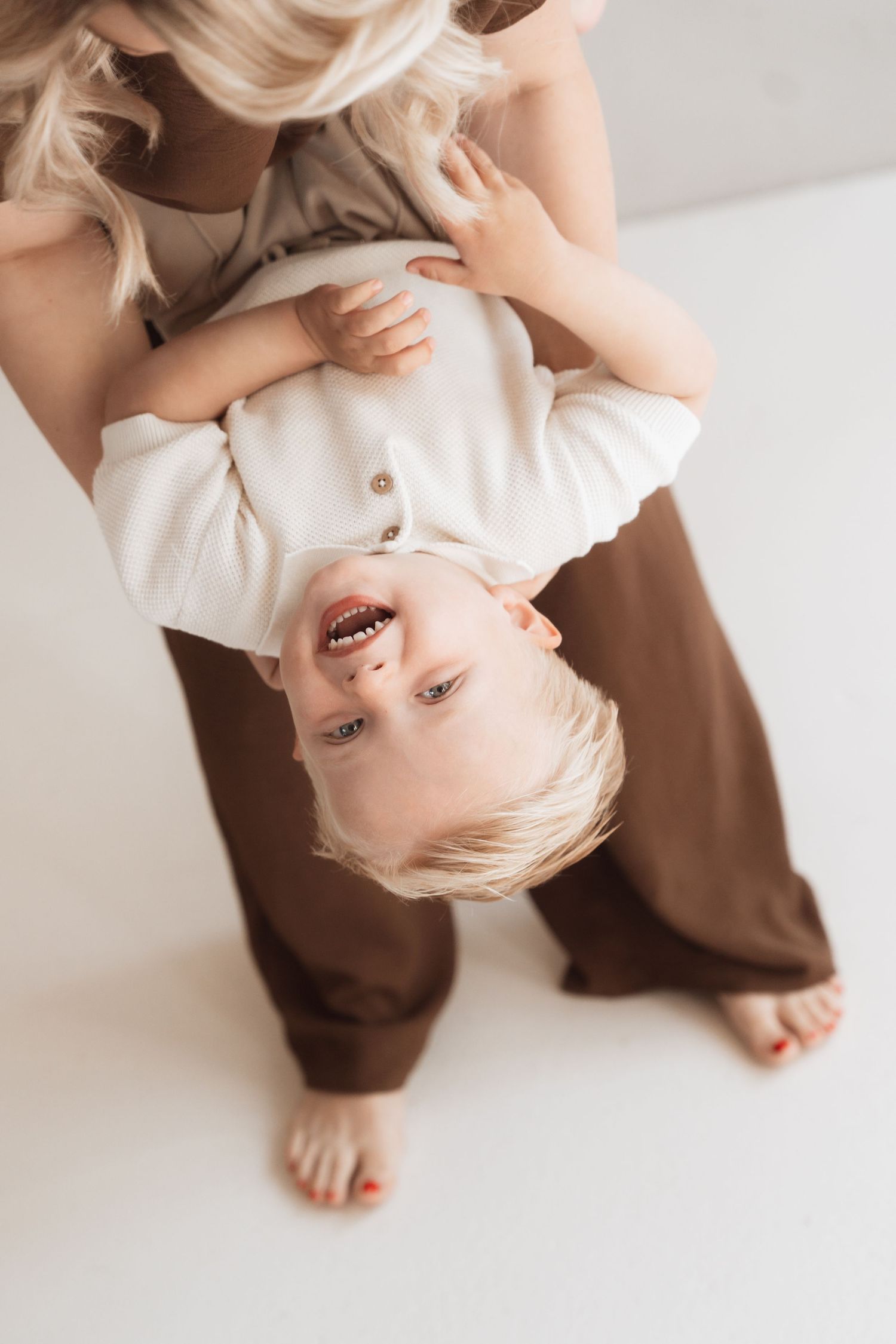 A laughing blonde toddler held upside down by a smiling blonde mother in a bright white studio setting.