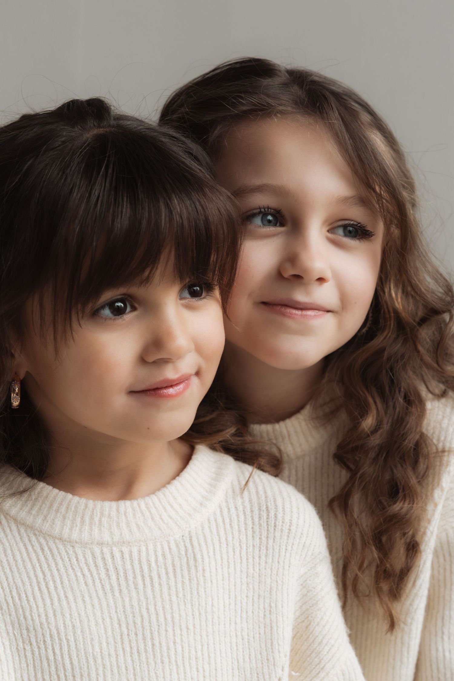 Two young girls with dark hair wearing white knit sweaters pose together smiling in a studio portrait.