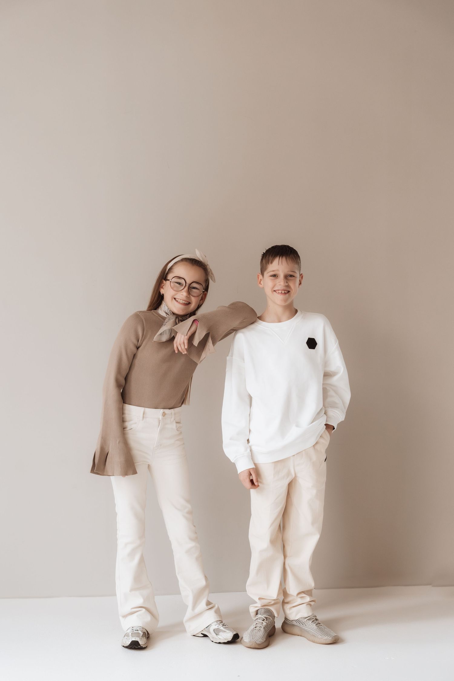 Two kids in neutral tones pose together, girl in brown sweater and glasses, boy in white sweatshirt, against beige wall.