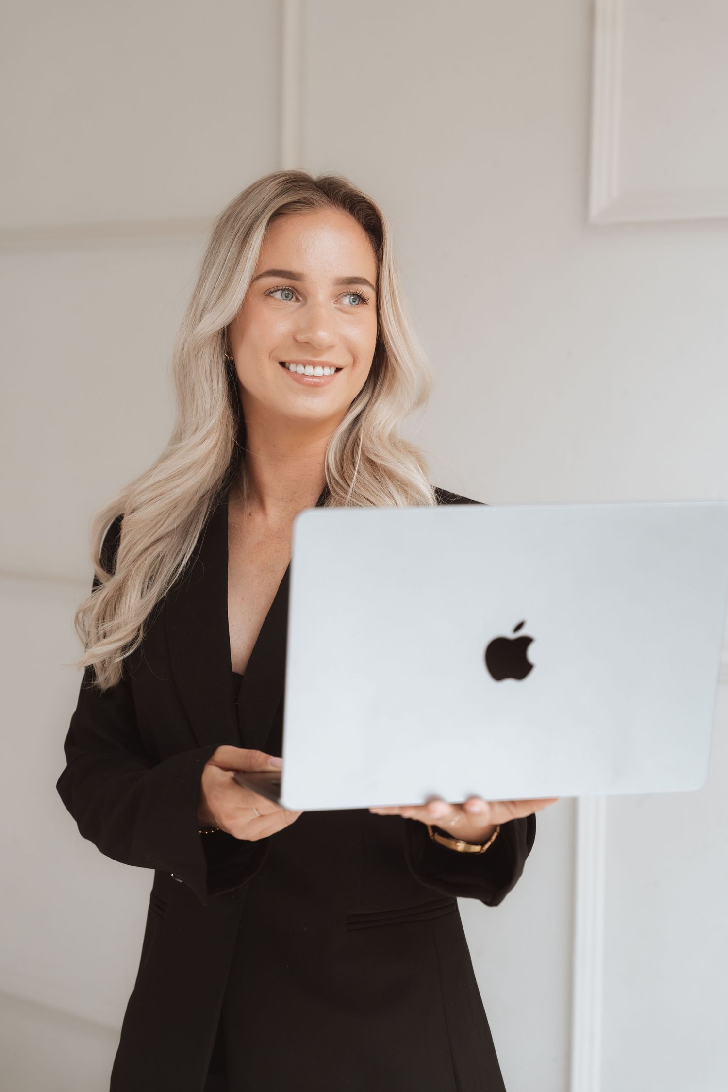 Smiling blonde woman in black blazer holding a silver Apple MacBook laptop in a bright modern office setting.