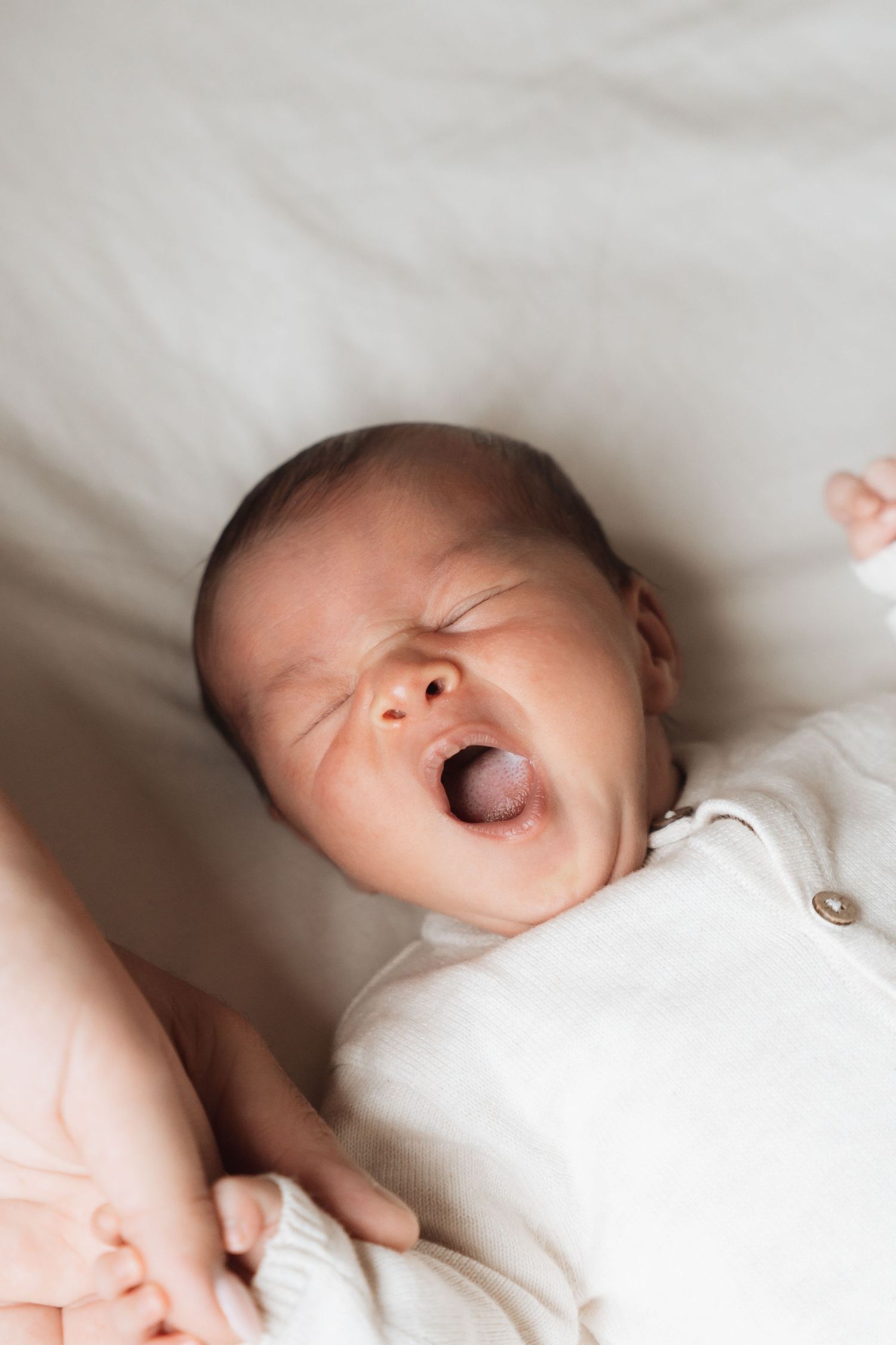 Newborn baby yawning while lying on a white blanket, wearing a white onesie, with eyes closed.