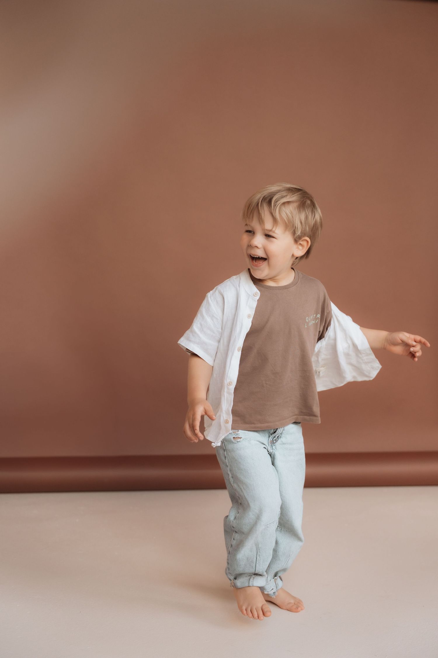 Happy toddler boy laughing and dancing barefoot, wearing light blue jeans and brown t-shirt with white shirt.