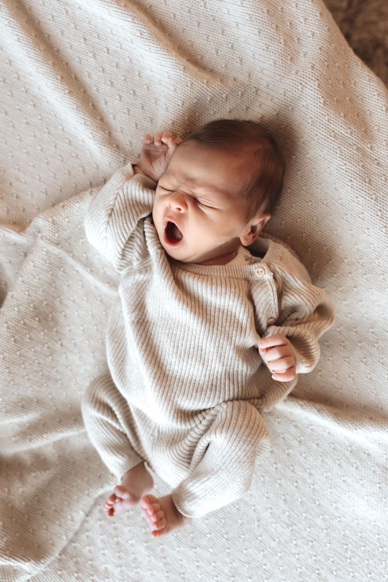 Baby in cream ribbed onesie yawning while lying on textured white bedding.