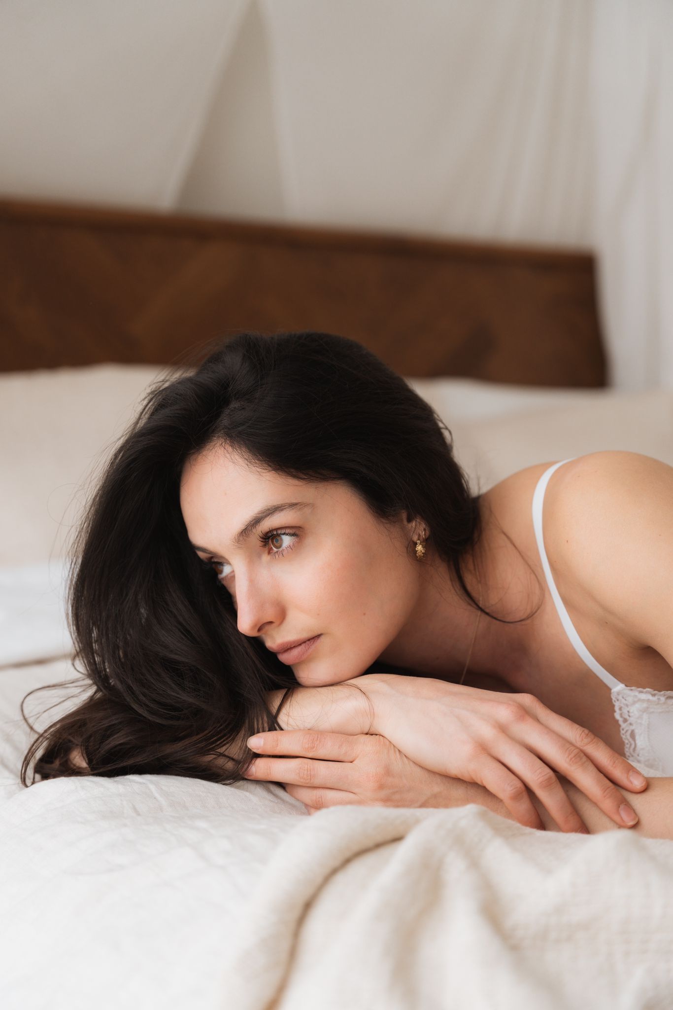 Woman with dark hair resting on white bedding in intimate portrait.