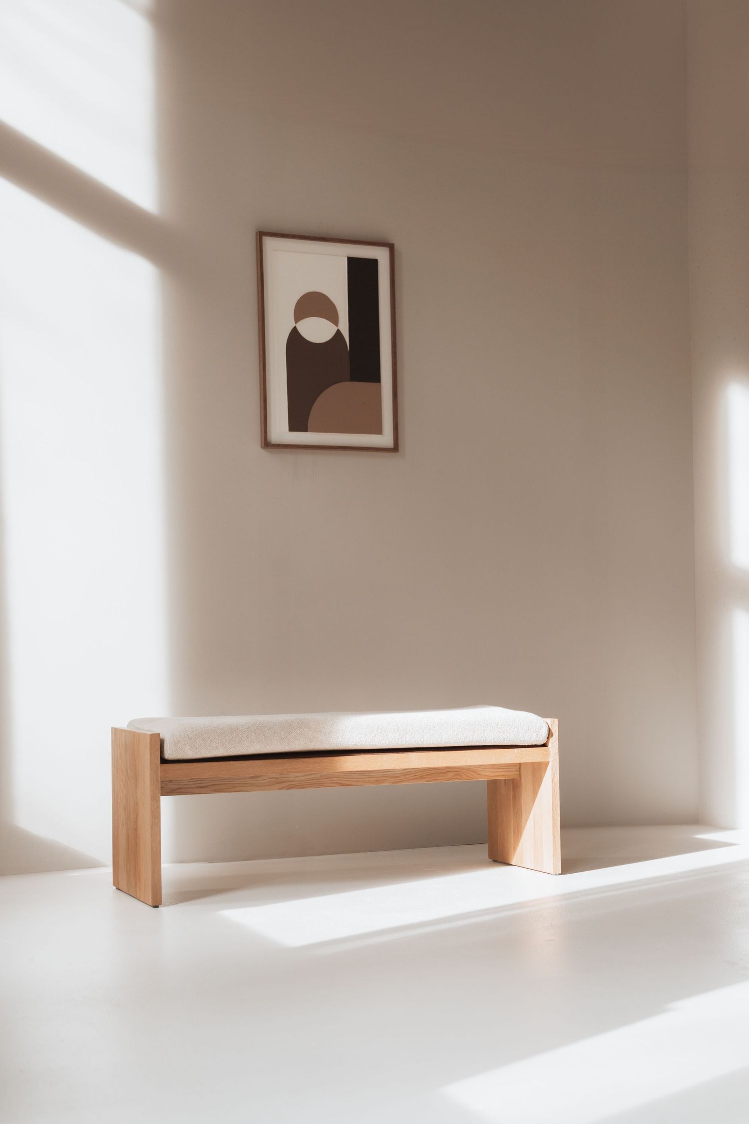 Minimalist wooden bed frame with white mattress casting dramatic shadows in a sunlit room with neutral walls.