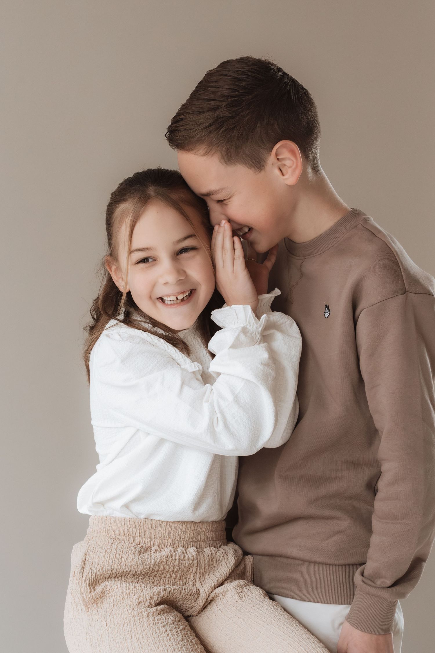 Two children sharing a secret, girl in white blouse laughing while boy in brown sweatshirt whispers in her ear.