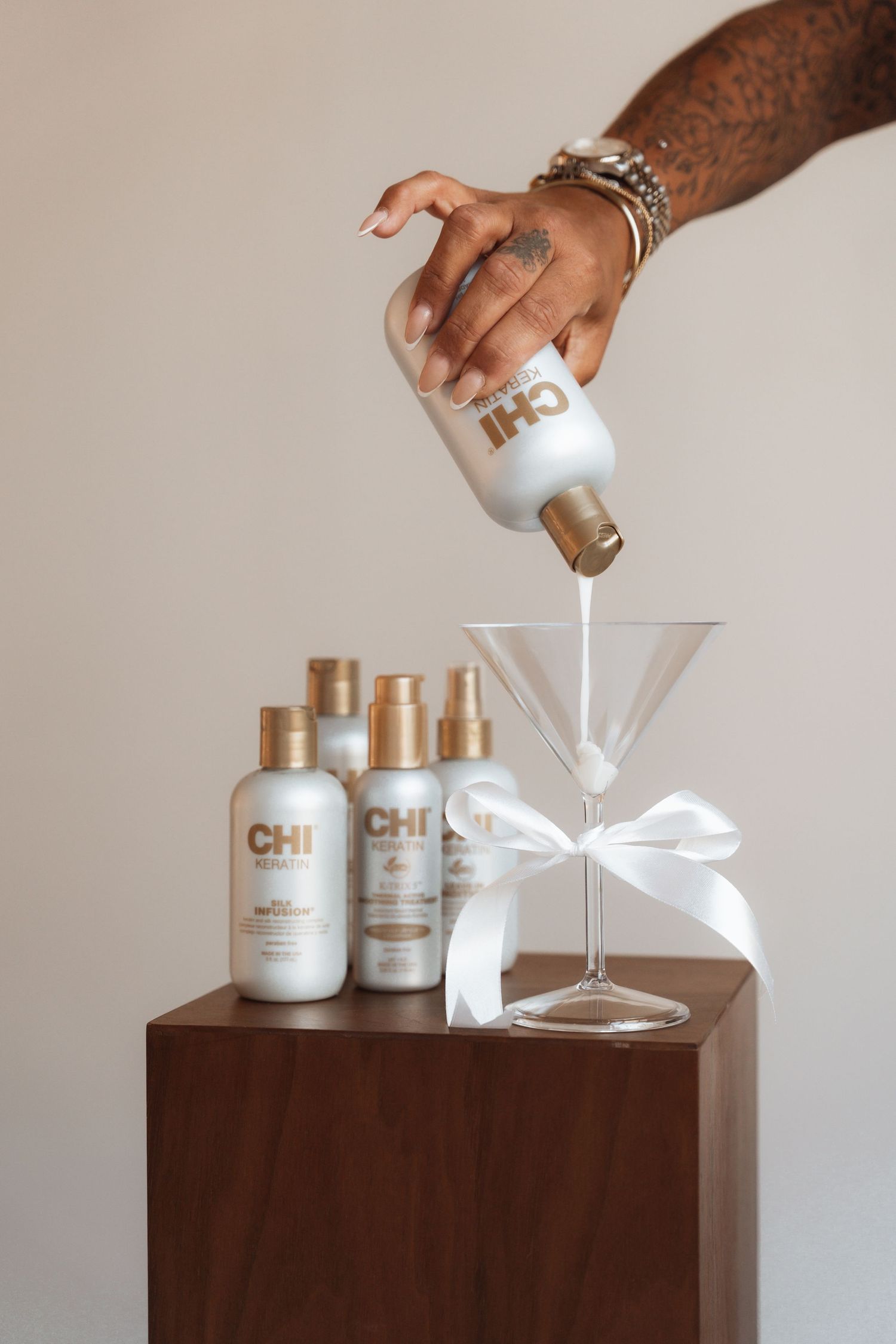 Tattooed hand pouring CHI haircare product into martini glass beside CHI bottles on wooden pedestal.