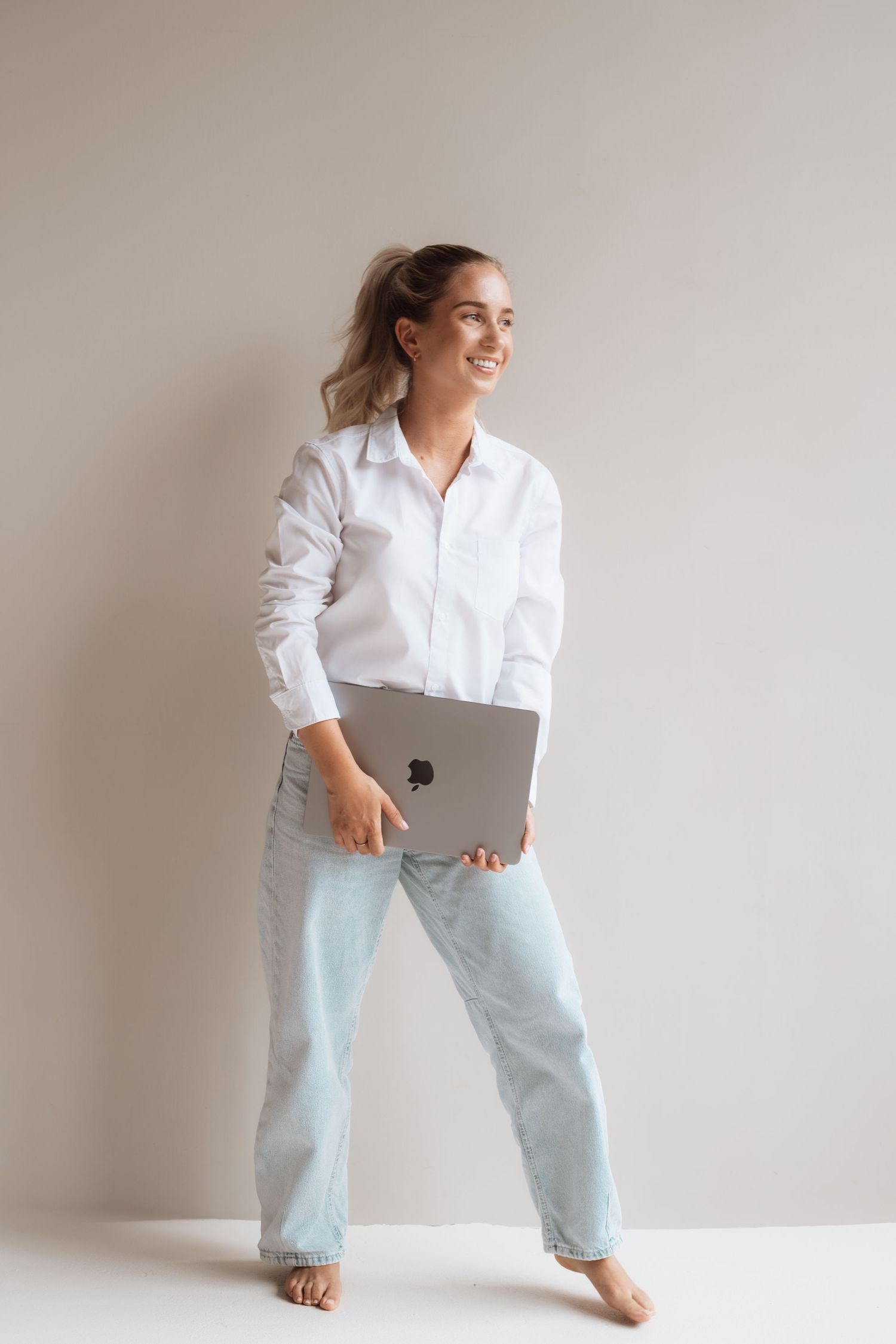 Smiling woman in white shirt and light jeans holding a laptop, standing against a neutral gray background.
