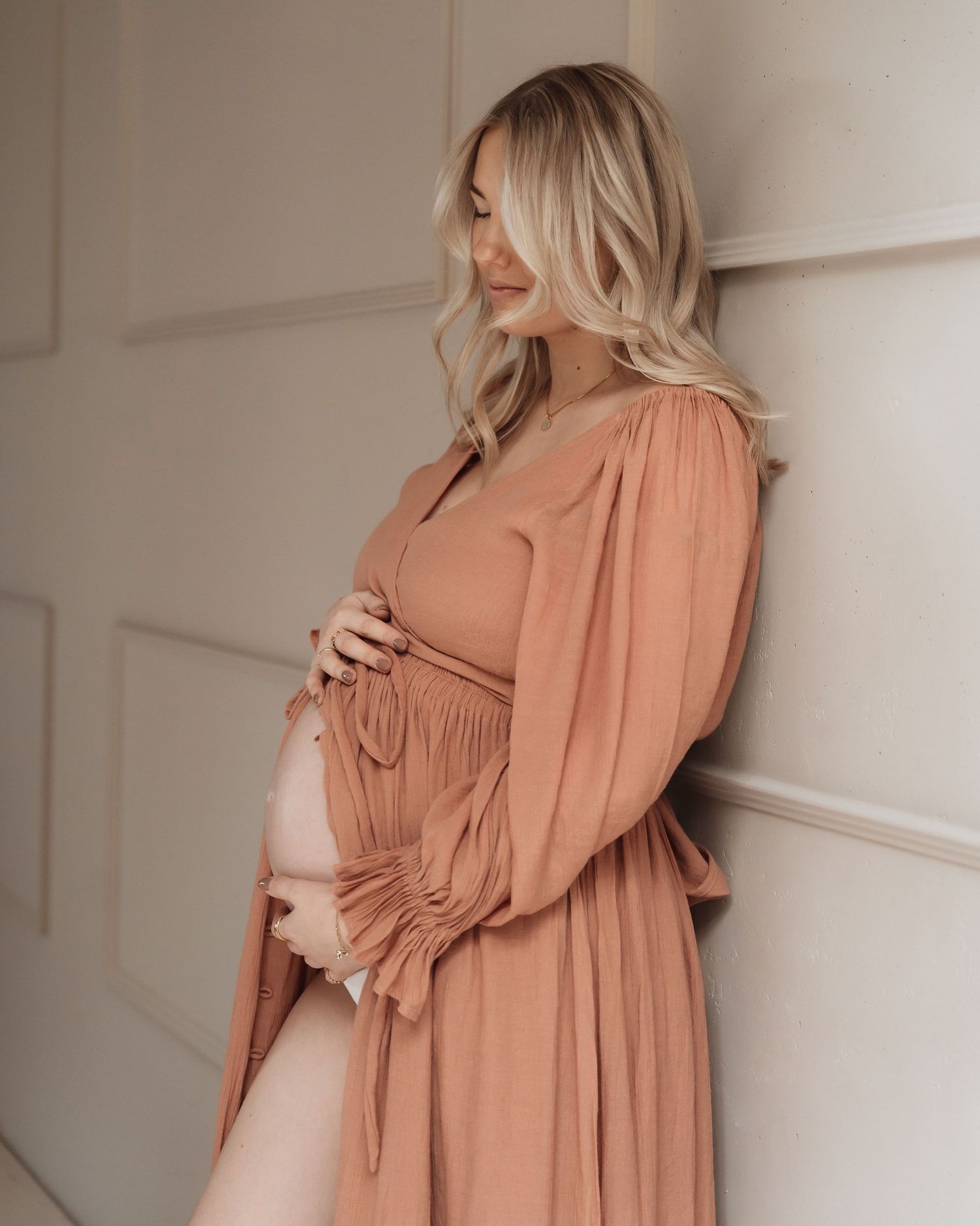 Woman in peach-colored dress holding her pregnant belly against white wall.