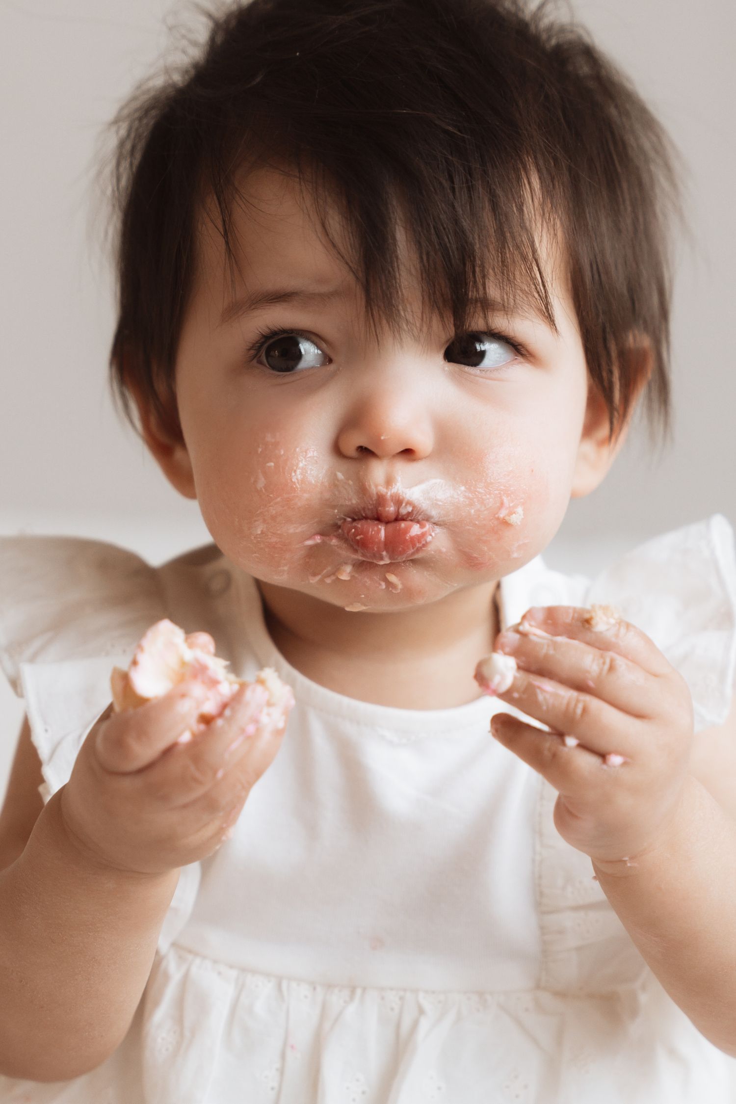 A messy baby girl in a white dress enjoys cake with frosting on her face and hands during her first birthday smash.