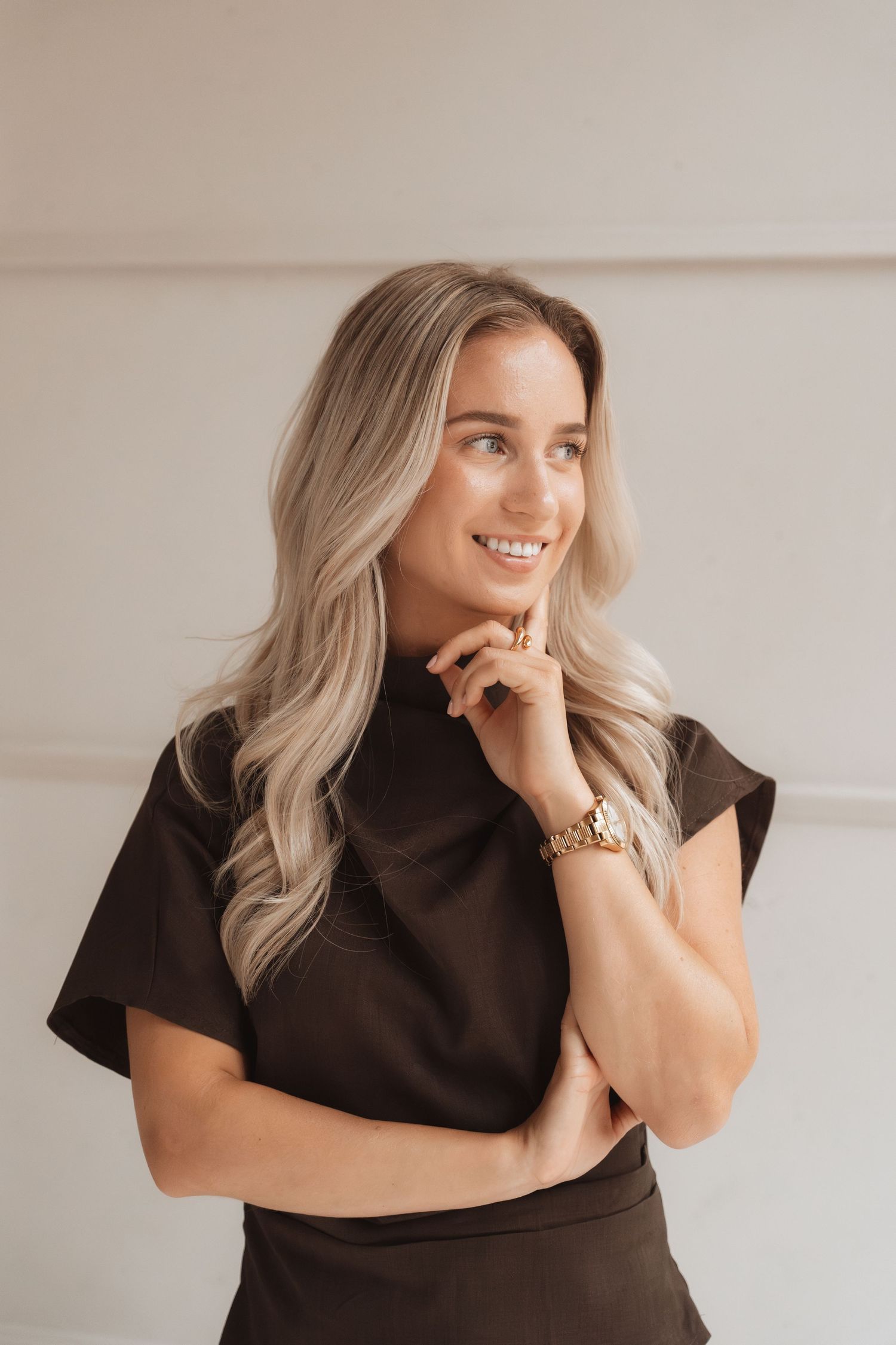 Smiling blonde woman in a dark top with a gold watch, posing thoughtfully with hand on chin against a light background.
