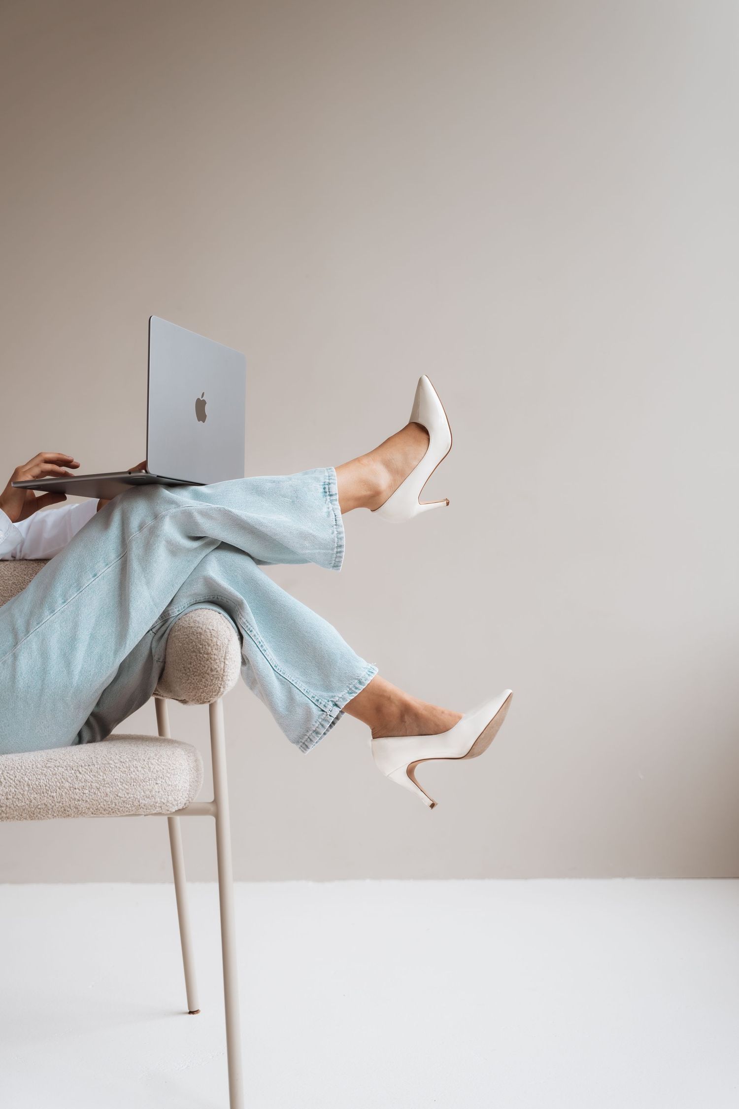 Woman in light blue jeans and white heels using a MacBook laptop while seated in a modern chair.