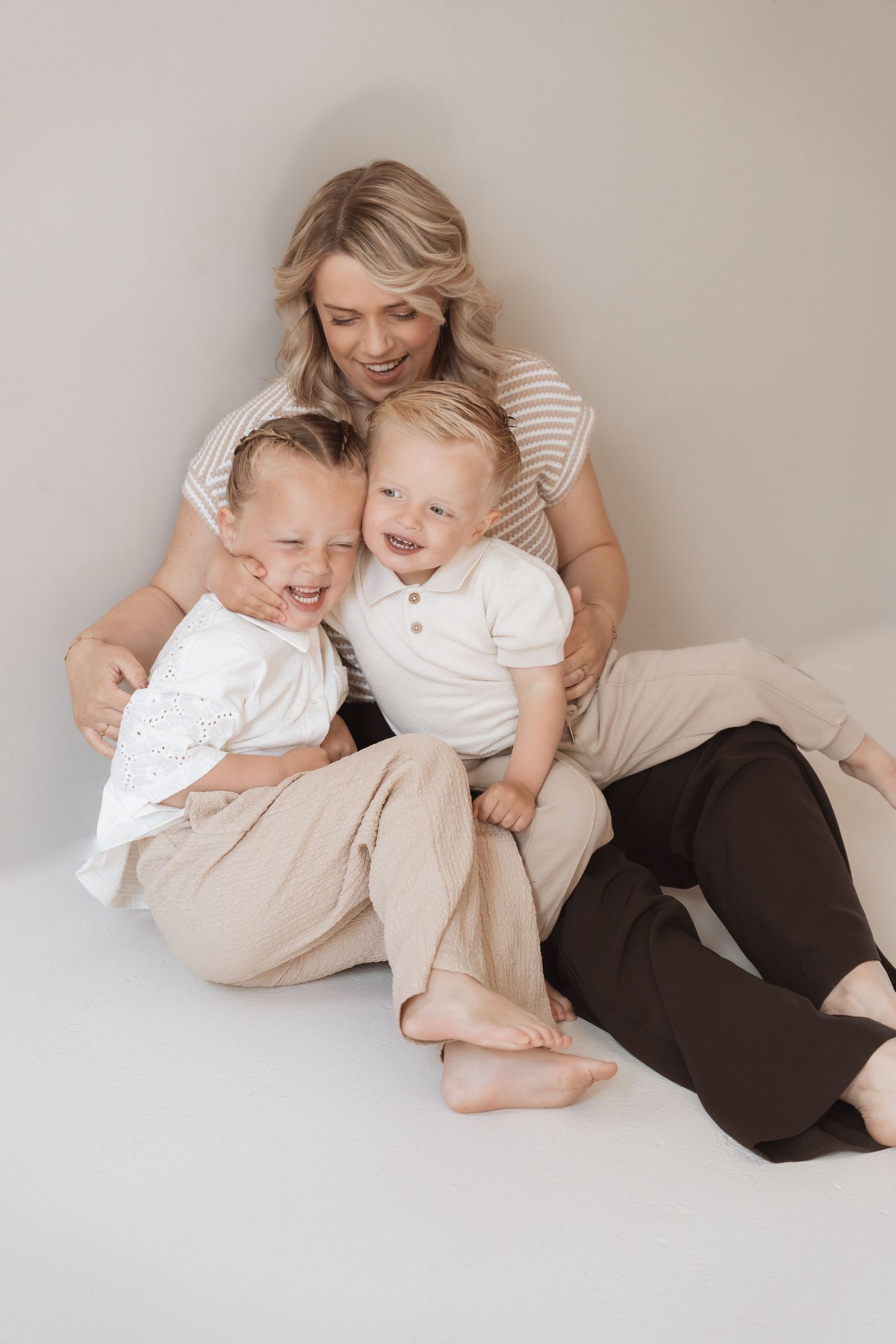 Smiling blonde mother sitting on floor hugging two laughing young boys in neutral tones for a family portrait.